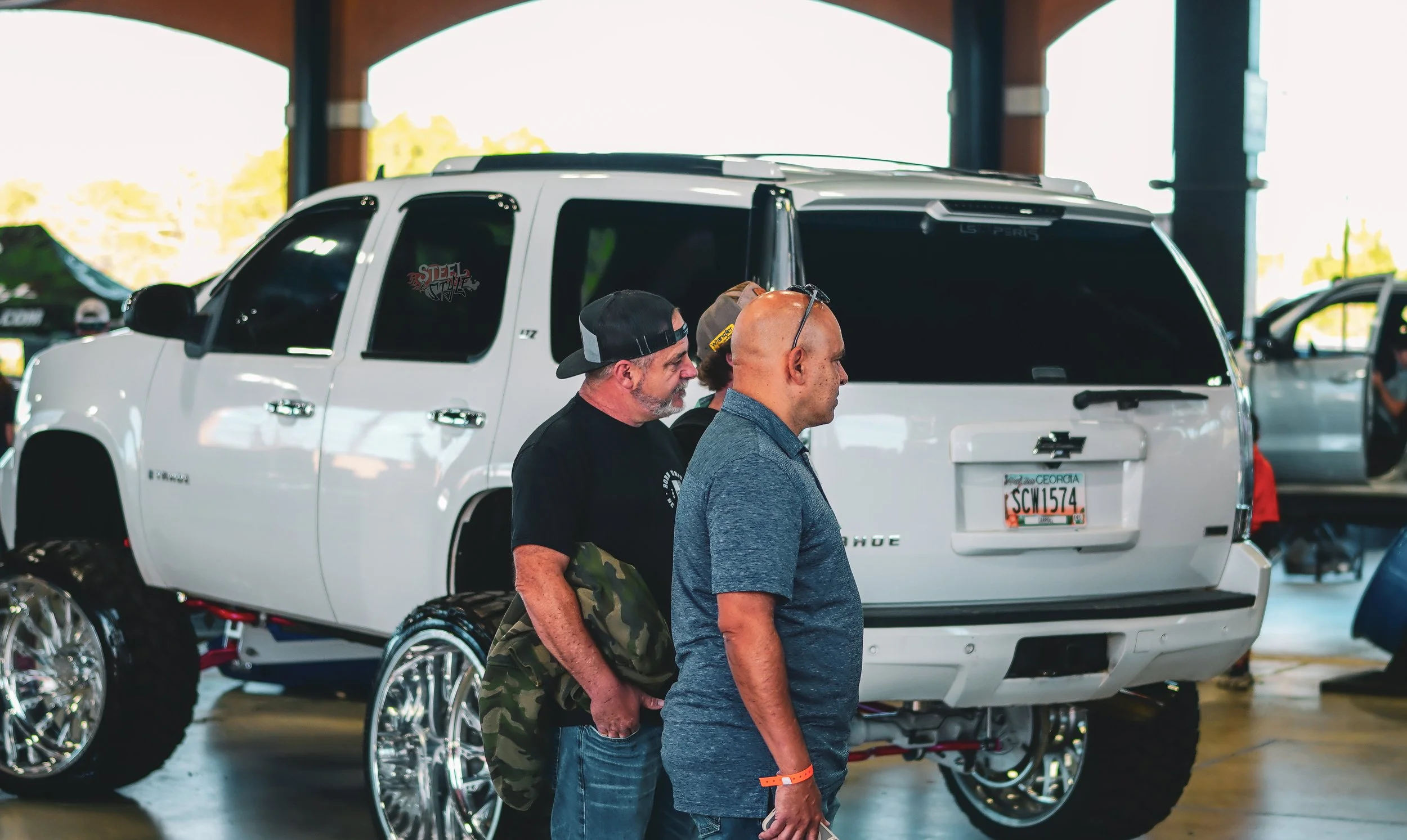 Three men standing in front of a white SUV with custom wheels inside a large indoor space, probably at a car show or event.