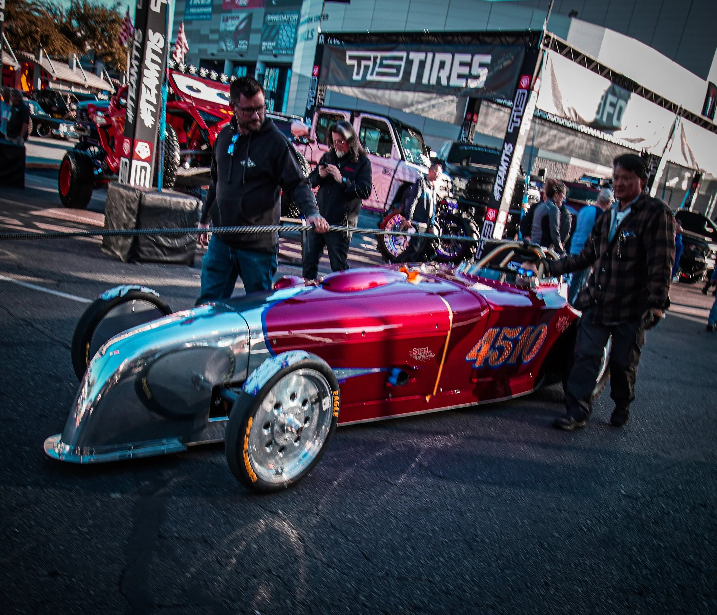 A vintage-style race car painted in red and silver with the number 450 on its side, being pushed by two men at an outdoor event. In the background, there are tents, motorcycles, and people walking around.