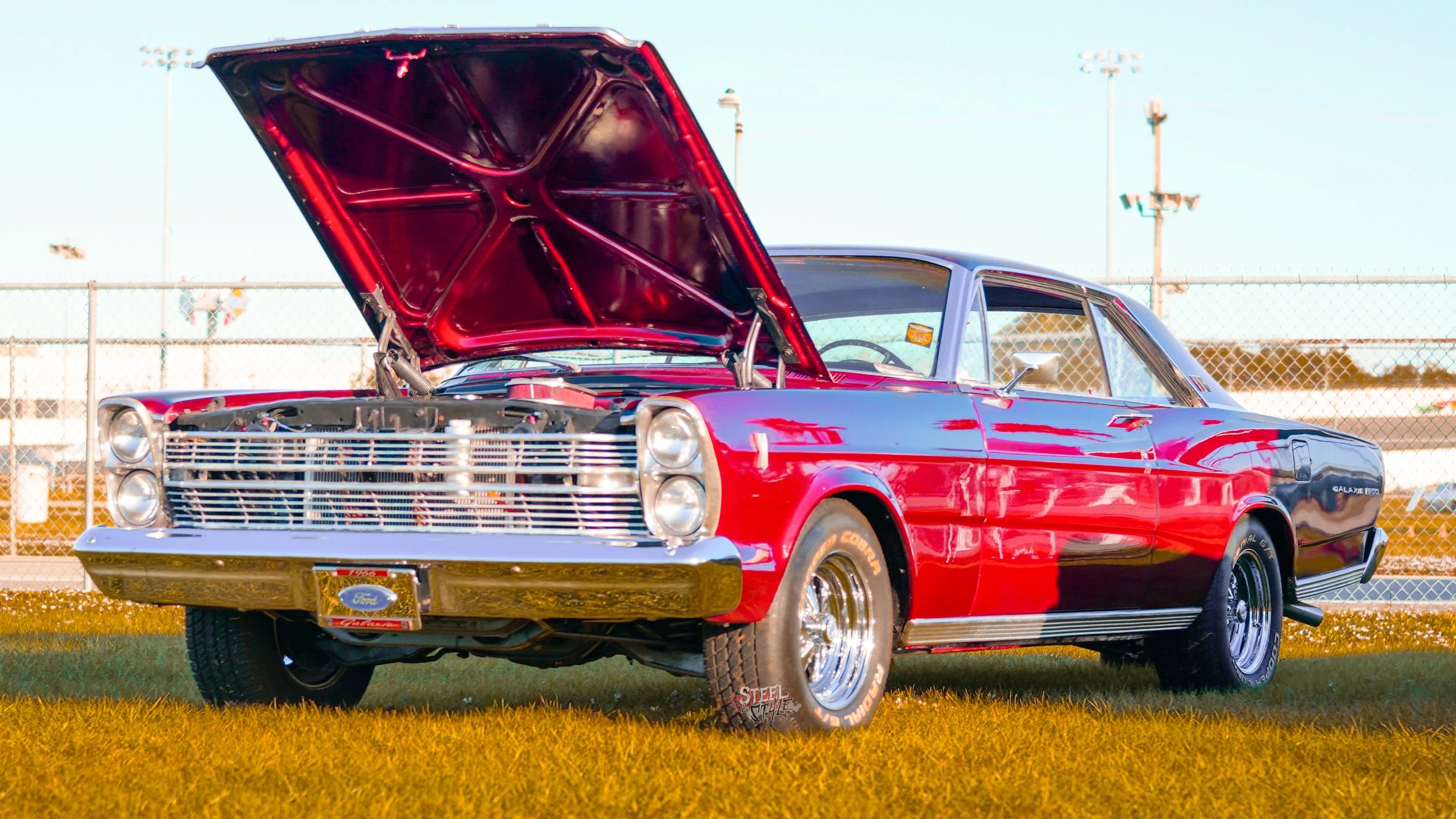 A red vintage Ford Galaxie 500 car with its hood open, parked on the grass at a racetrack, with chain-link fencing and light poles in the background.