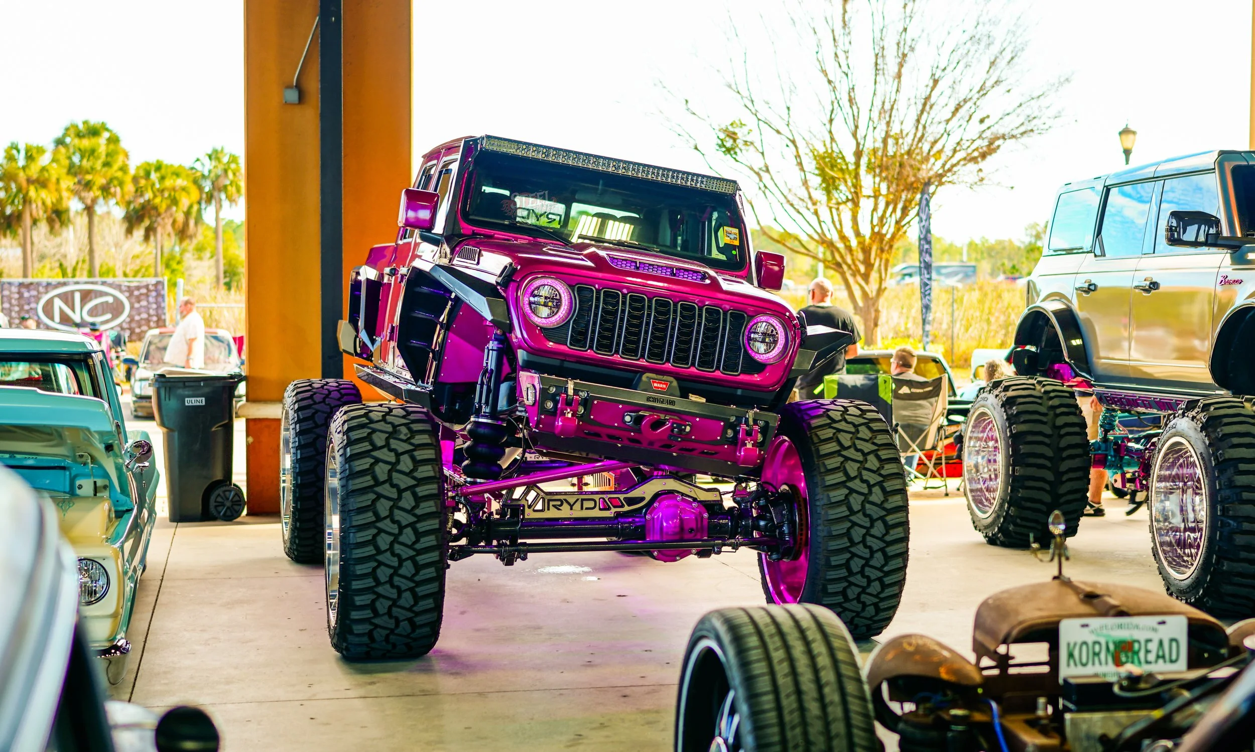 A highly customized purple off-road Jeep with oversized tires is prominently displayed at an automotive show, with other cars and people in the background.
