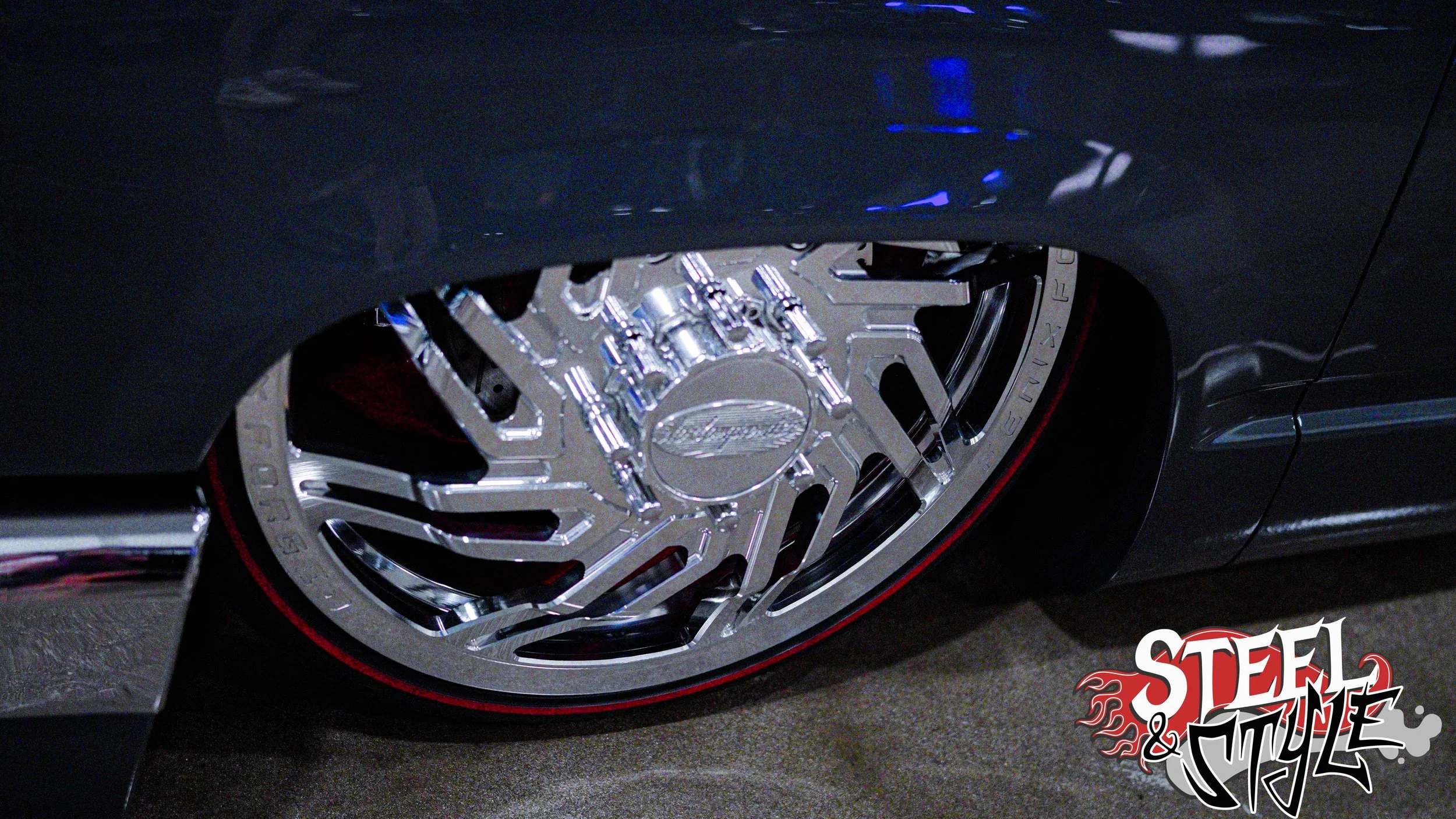 Close-up of a shiny, chrome custom wheel on a dark-colored car, with a red accent line on the tire. The wheel has an intricate, geometric design with a center cap. The image has a logo dealership or brand name 'Steel & Style' in the bottom right corn