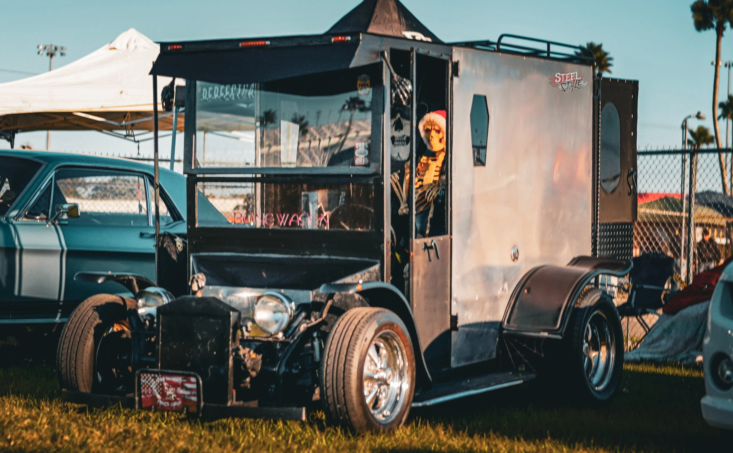 A vintage hot rod car with a trailer attached, parked on grass surrounded by tents and palm trees, at an outdoor event during sunset.