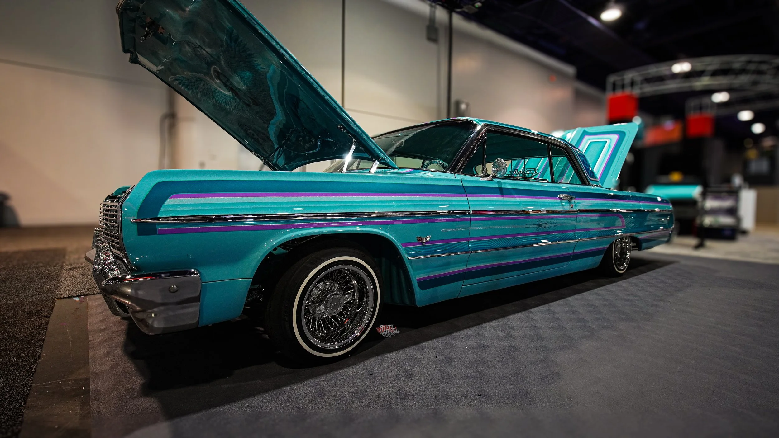 A vintage lowrider car with turquoise paint and colorful stripe accents, displayed at an indoor car show with its hood and trunk open.