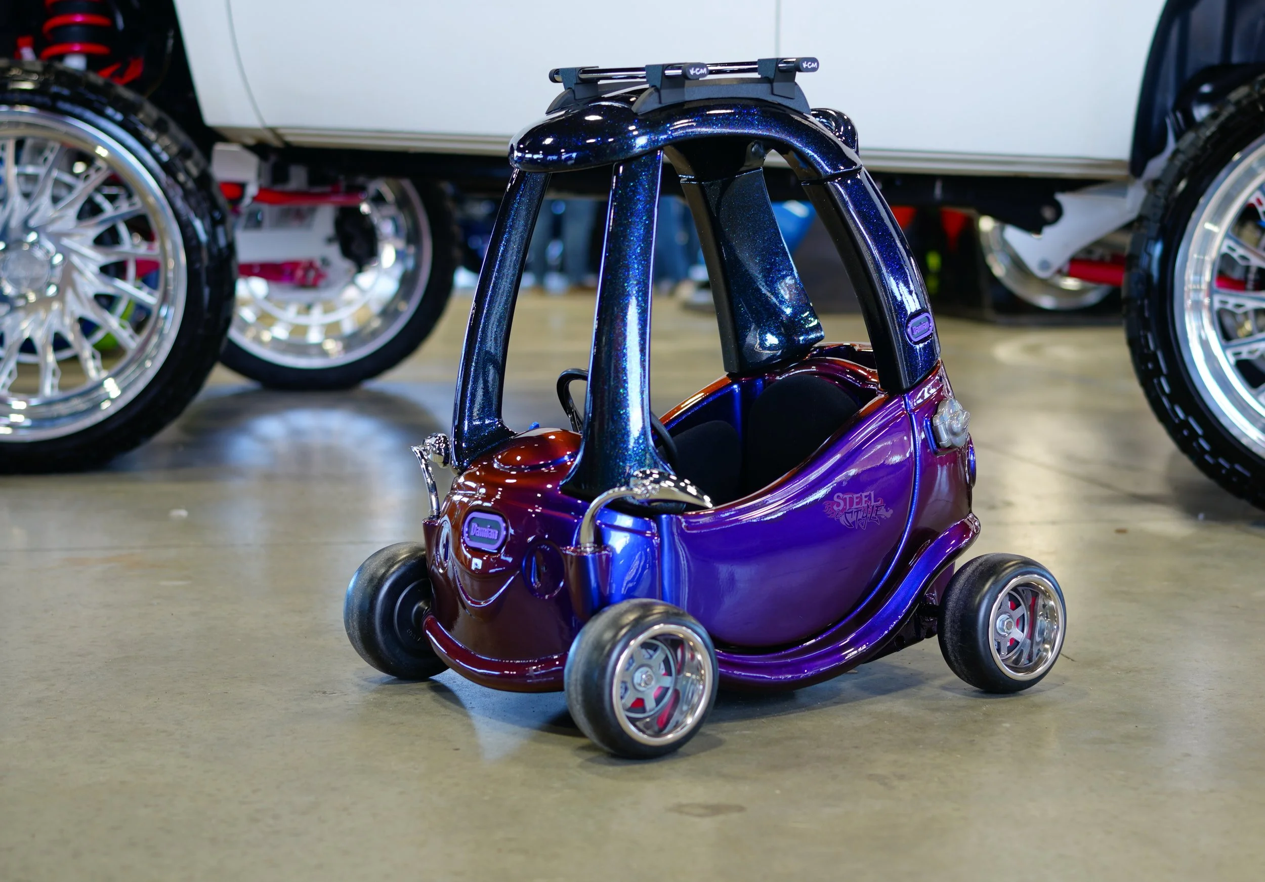 A miniature toy car modeled like a vintage bubble car in metallic purple with a black roof and silver wheels, situated on a showroom floor with other vehicles and large tires in the background.