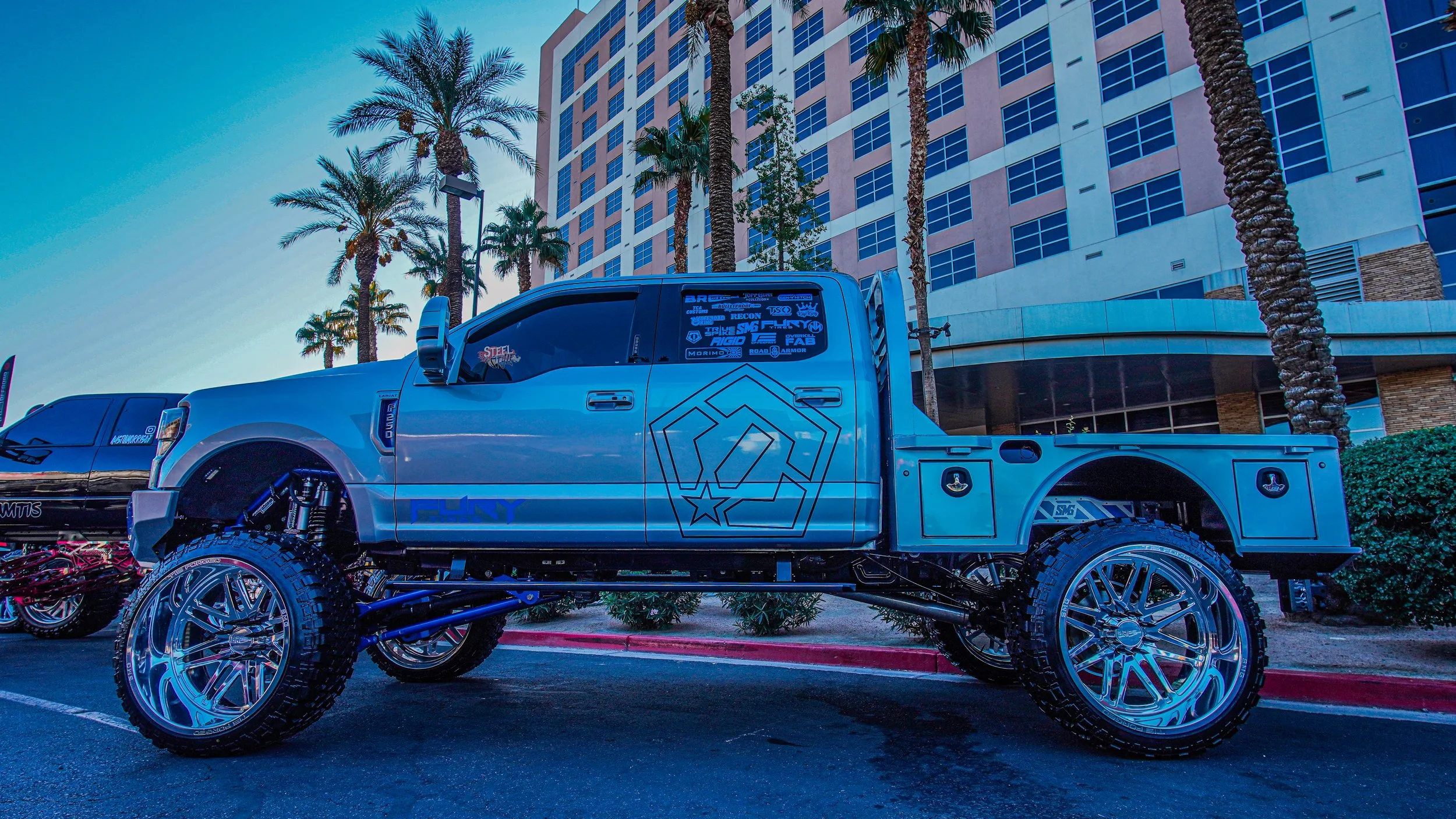 A customized lifted pickup truck with large chrome wheels and off-road tires, parked in front of a high-rise building with palm trees.
