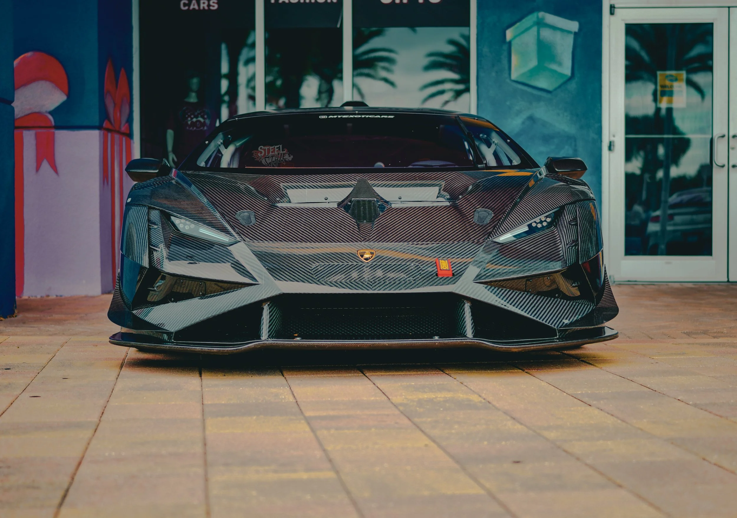 A high-performance sports car with a black carbon fiber exterior parked in front of a storefront with signage that reads 'CARS,' 'FASHION,' and 'SHOP.' The car is facing forward, showcasing its sleek design and aggressive front splitter.