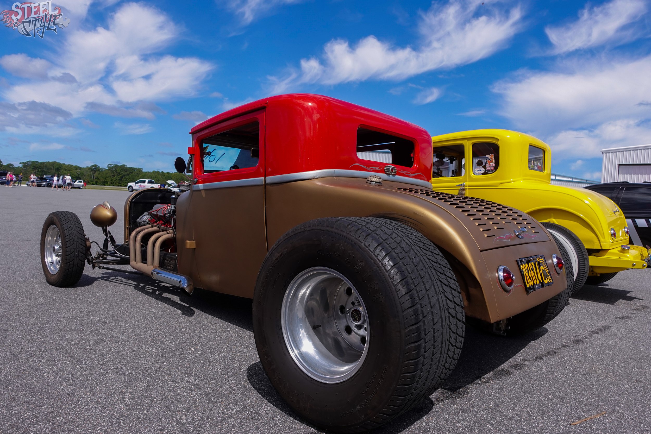 A vintage race car with a red and tan body parked on a tarmac lot under a partly cloudy sky, with a yellow car and people in the background.