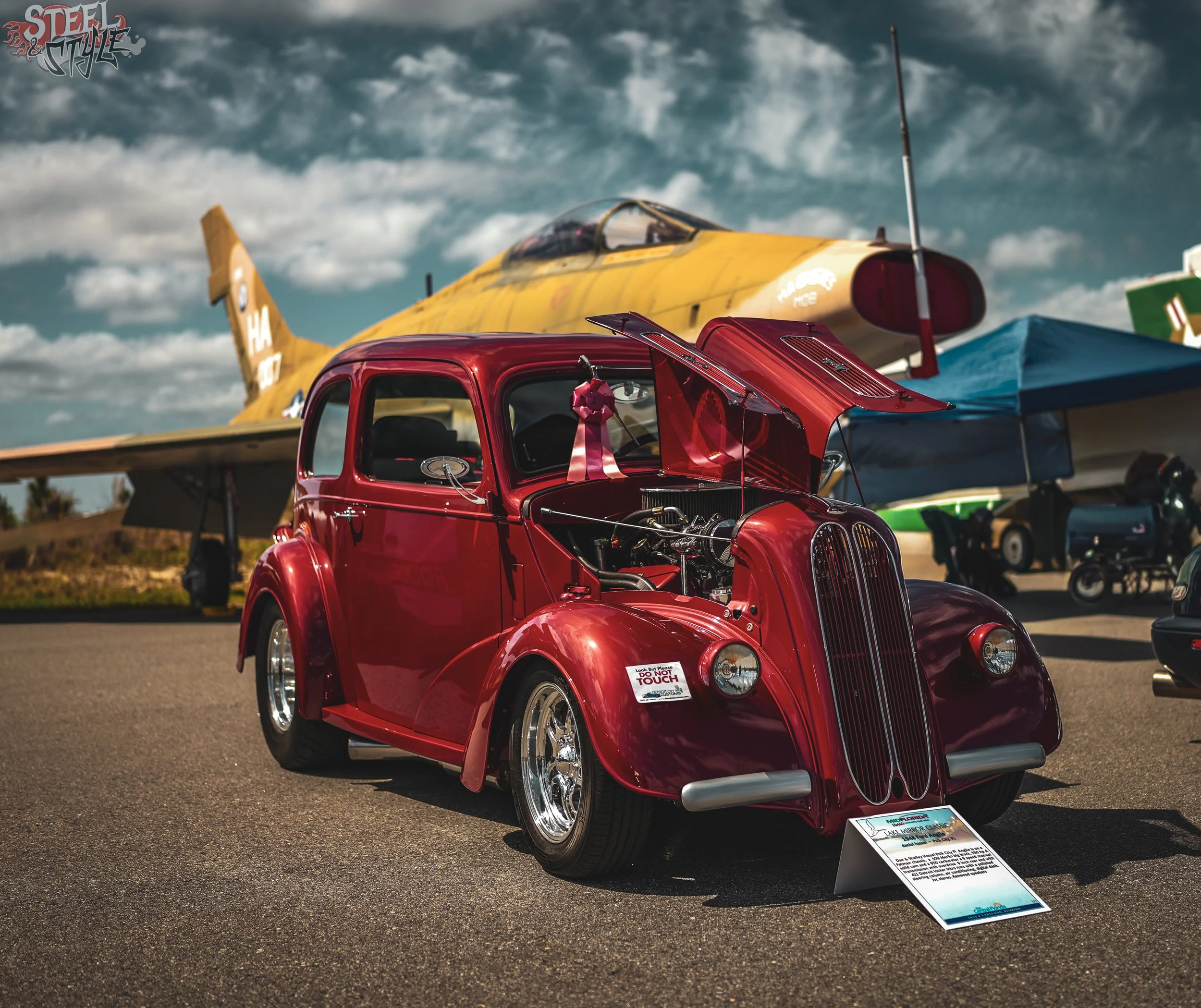A red vintage car with an open hood and a pink ribbon tied around the side mirror, displayed at an outdoor car show with a yellow airplane and a cloudy sky in the background.