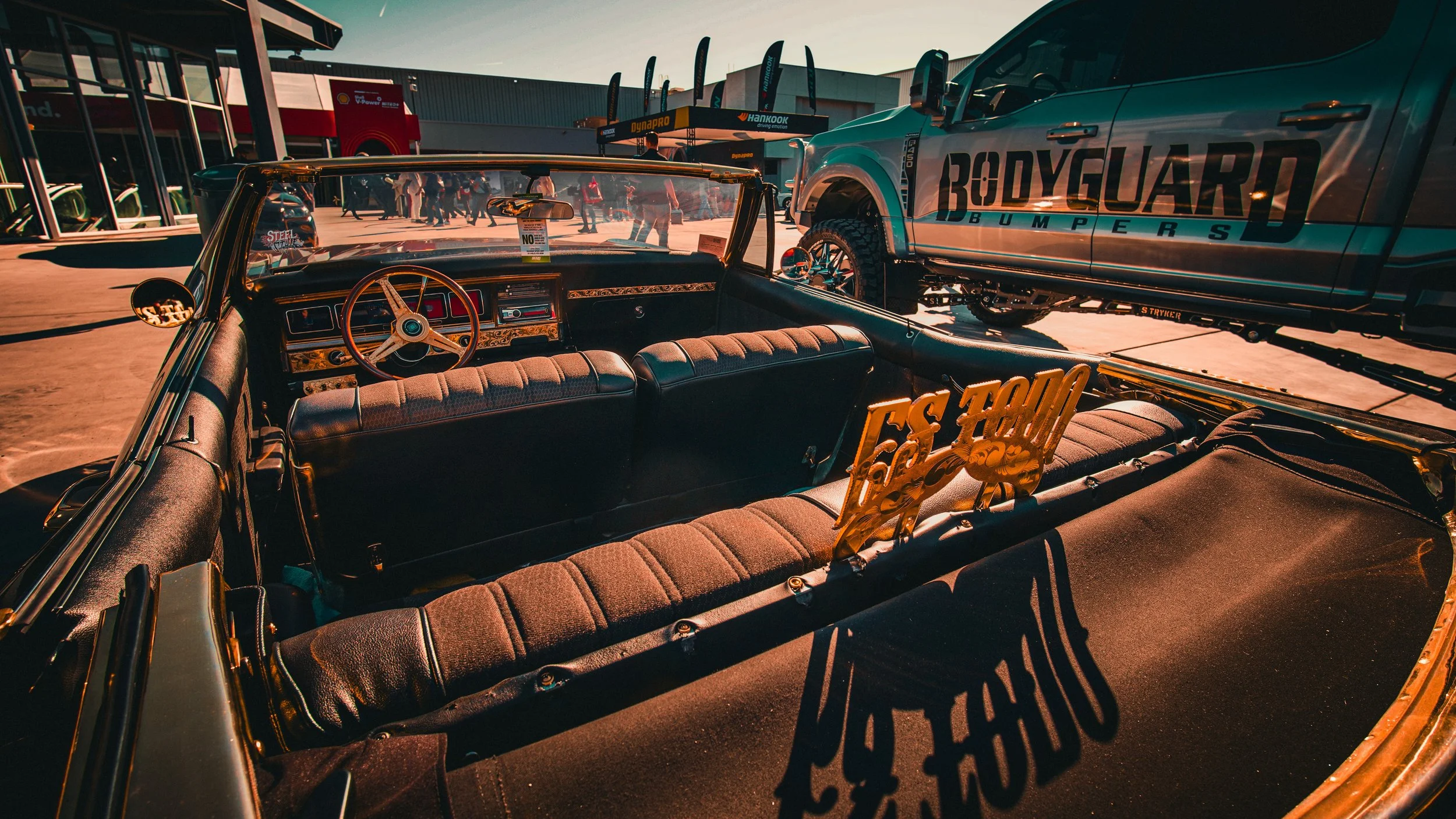 Vintage convertible car with a wooden steering wheel and luxurious interior, parked at an outdoor event with a large truck and crowds in the background during sunset.