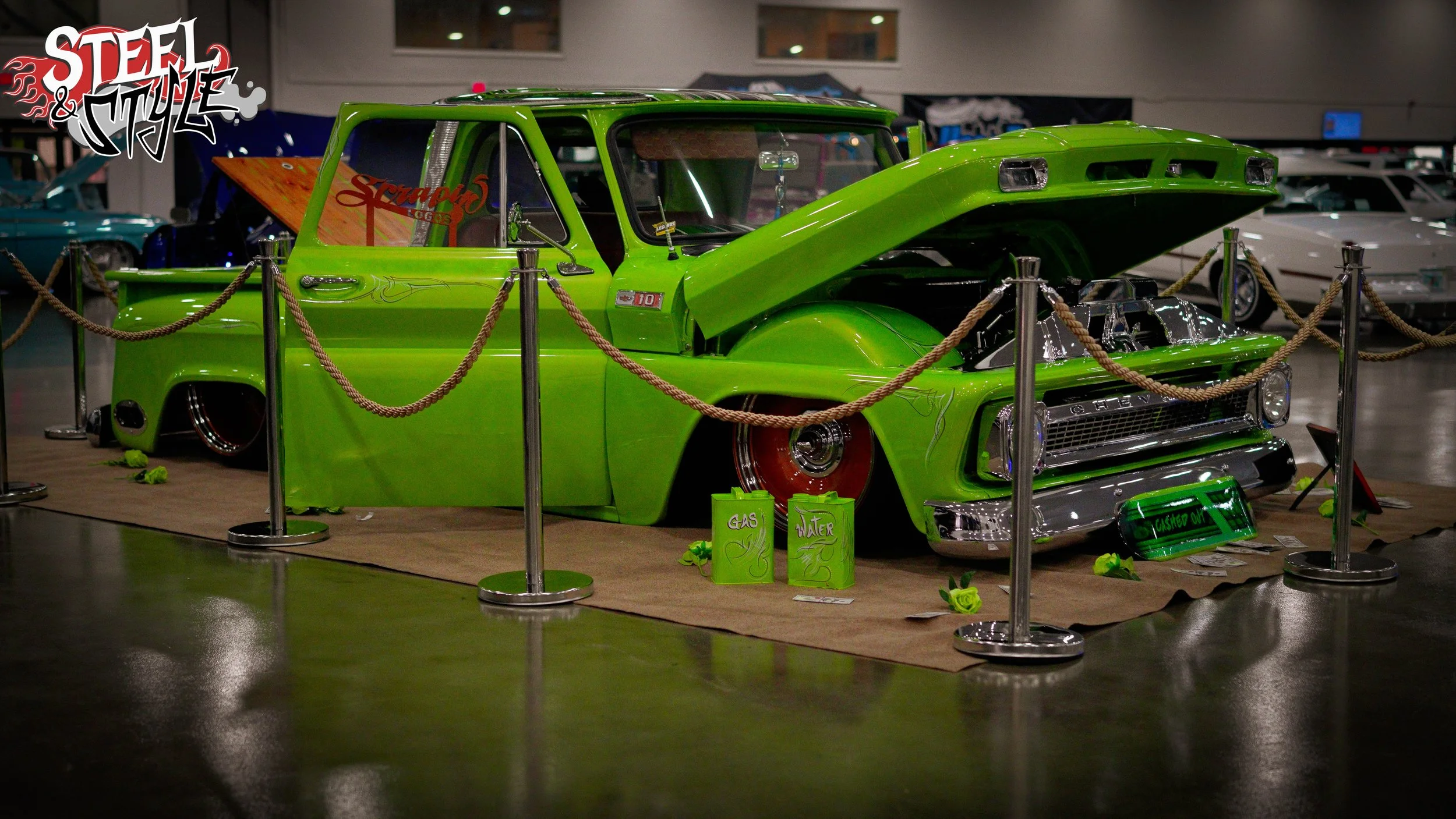A bright green customized vintage pickup truck on display at an indoor car show, surrounded by a rope barrier, with the hood open and various signs and decorative items around it.