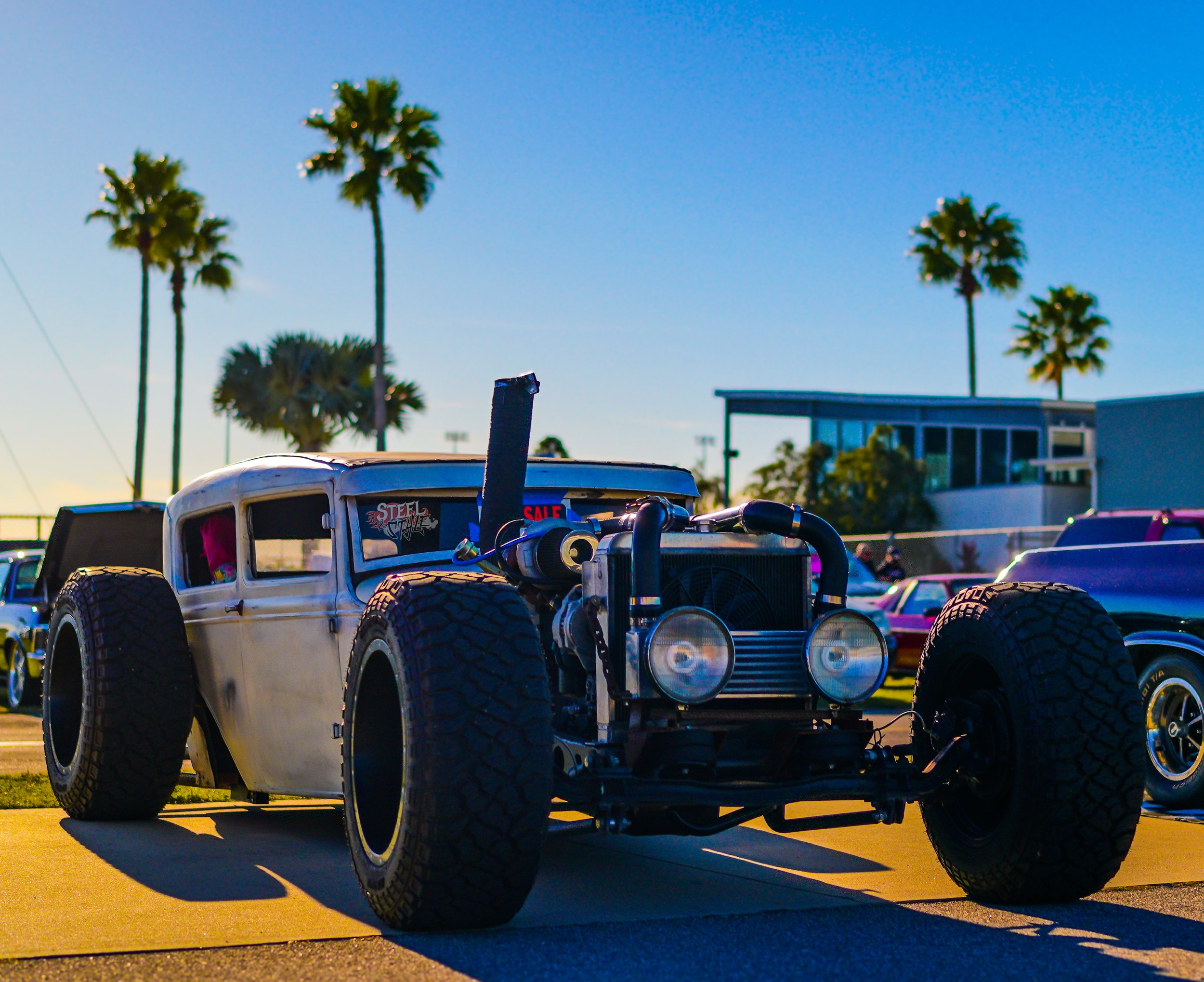 A vintage-style race car with large tires and a partially exposed engine, parked outdoors under a clear blue sky with palm trees in the background.