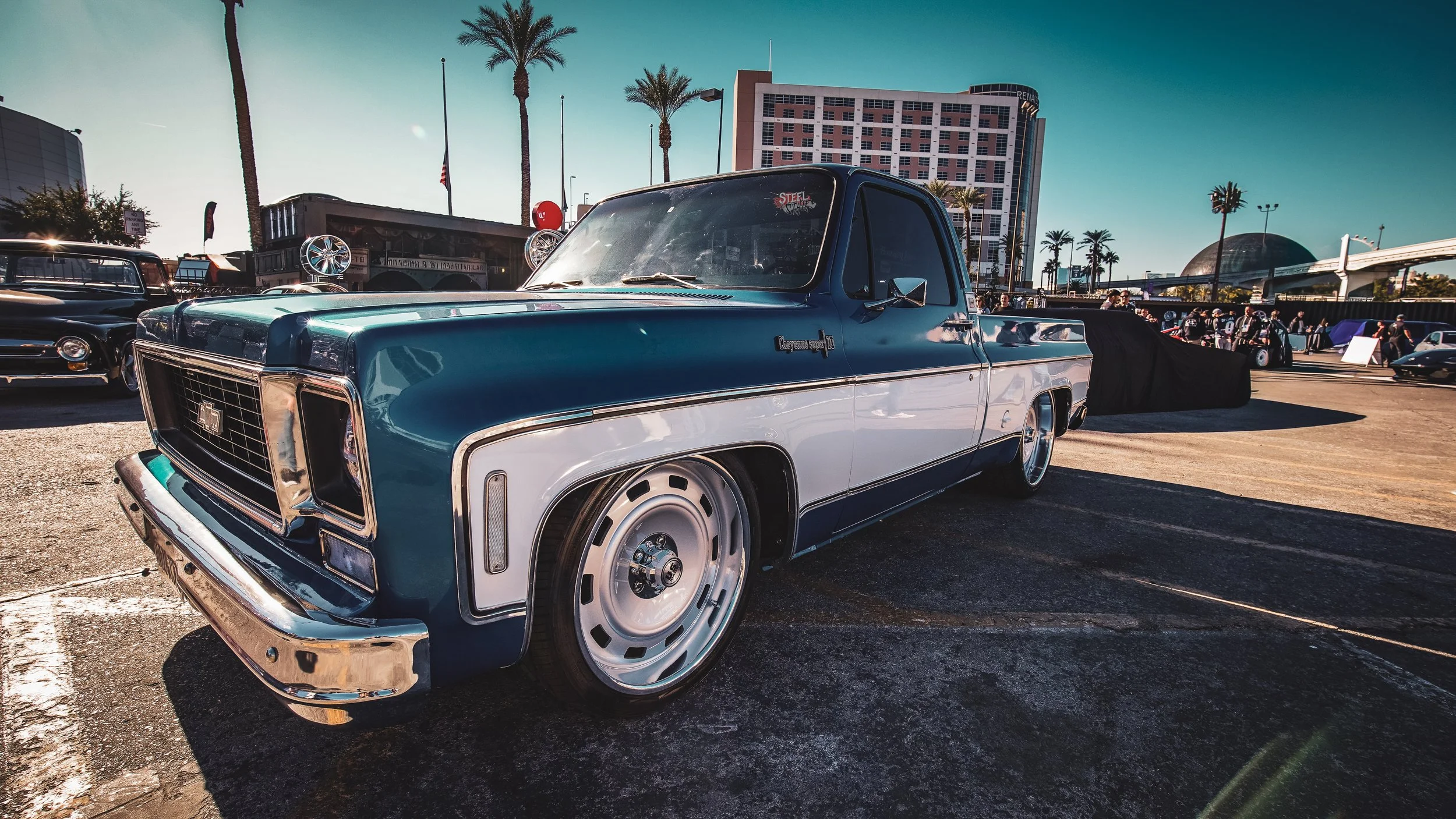A classic blue and white pickup truck on display at a car show, with other vintage cars and palm trees visible in the background.