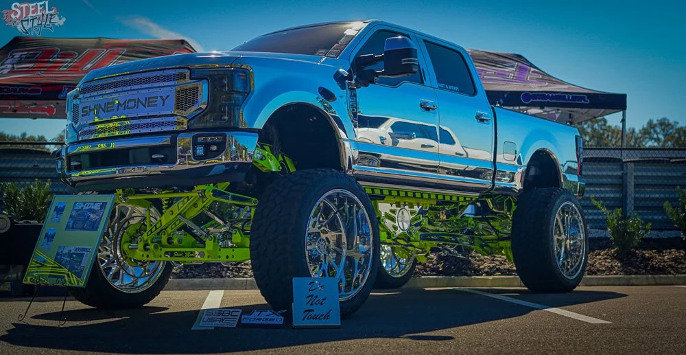 A modified large pickup truck with oversized green suspension and shiny chrome wheels on display at an outdoor event, with signs and banners in the background.