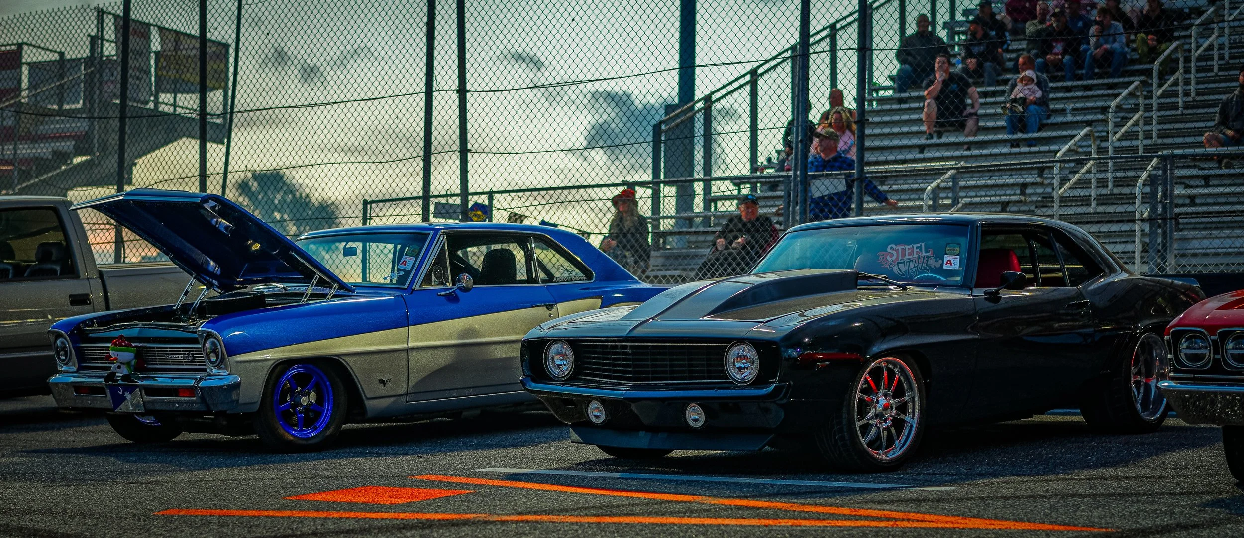 Two classic cars parked on a racing track, one blue with a cream stripe and the other black, with spectators sitting on bleachers in the background.