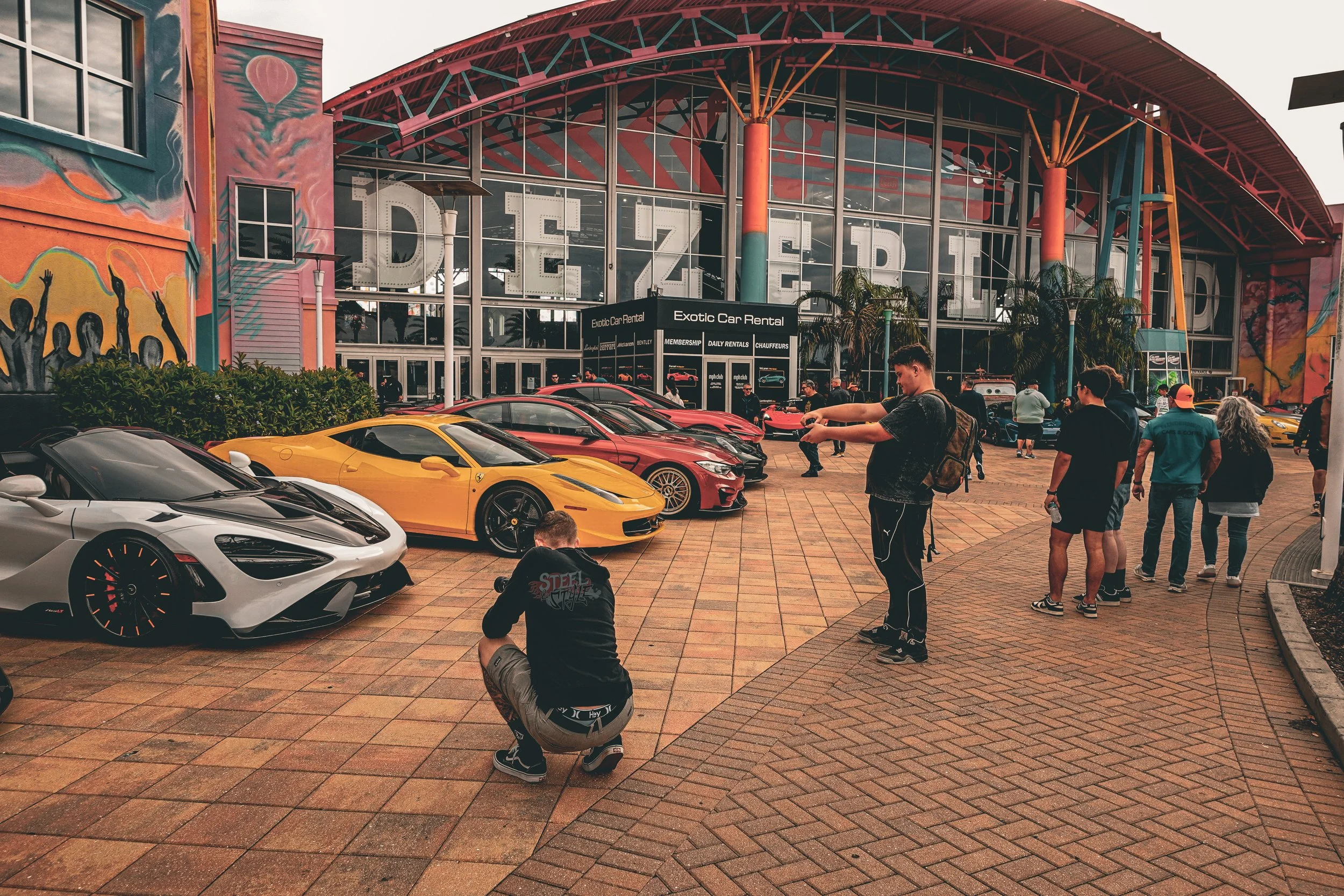 Lineup of luxury sports cars including a silver McLaren, a yellow Ferrari, and a red Tesla outside a colorful building with large glass windows and a sign that reads "Dezerland". People are walking, taking photos, and admiring the cars.