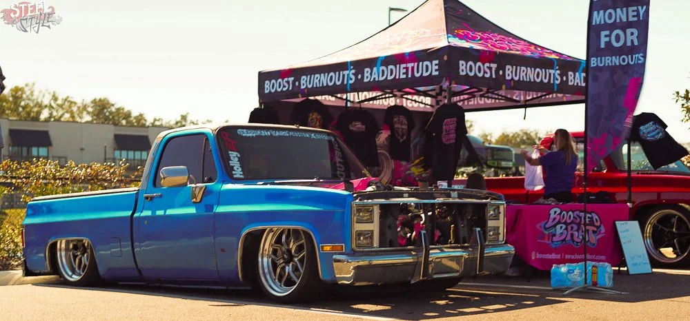 A blue lowered pickup truck parked next to a booth with a black and pink canopy that has various promotional signs and merchandise, at an outdoor event.