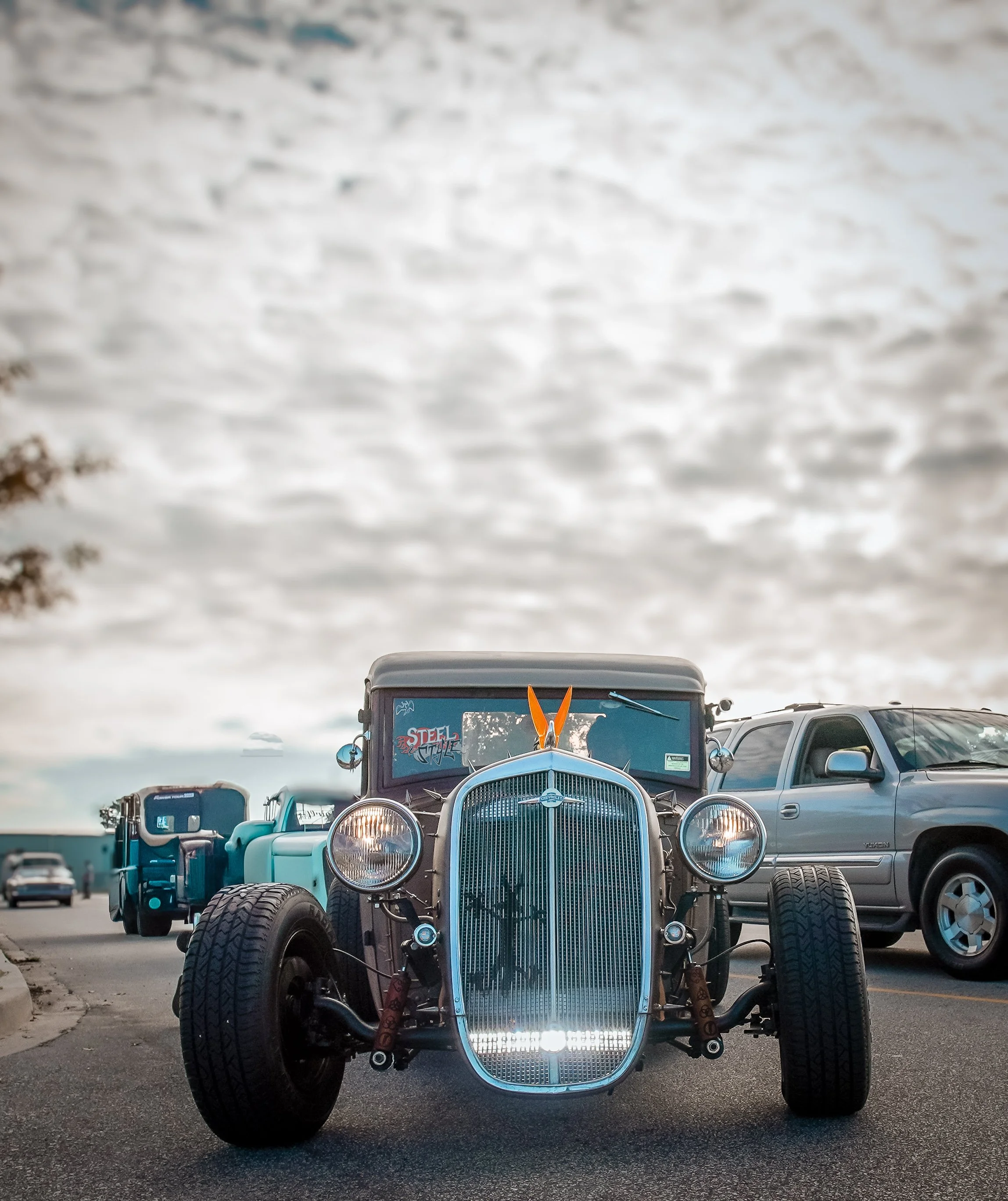 Vintage hot rod car parked on a street with other vehicles and cloudy sky in the background.