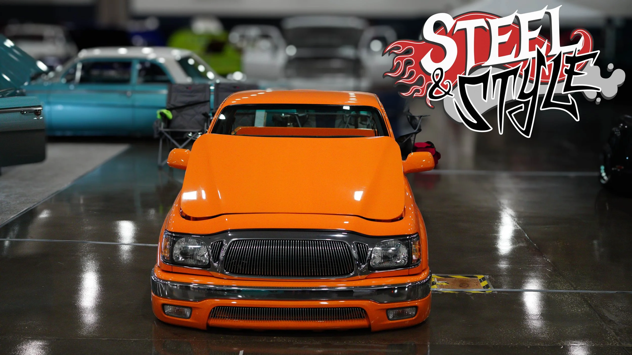 Orange custom car on display at a car show with other vehicles in the background and a "Steel & Style" logo in the top right corner.