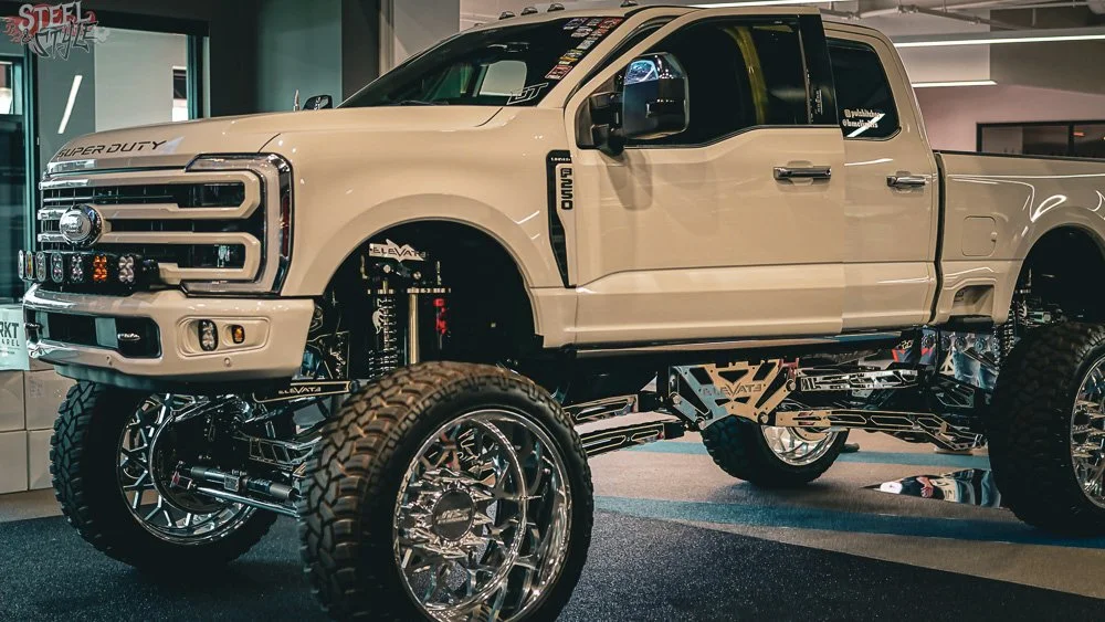 Custom lifted Ford Super Duty pickup truck with chrome rims, off-road tires, and a beige paint job, displayed indoors at an automotive show.