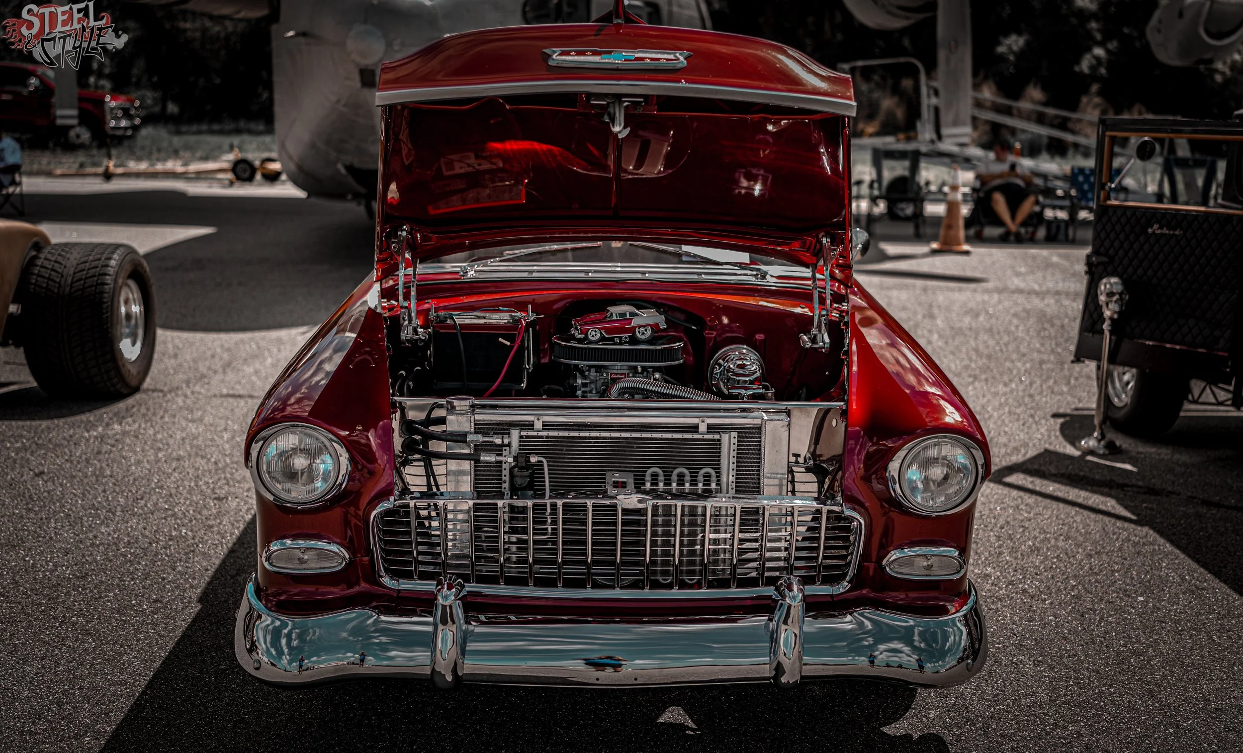 Red vintage Chevrolet car with its hood open, showcasing the engine at an outdoor car show on a sunny day.