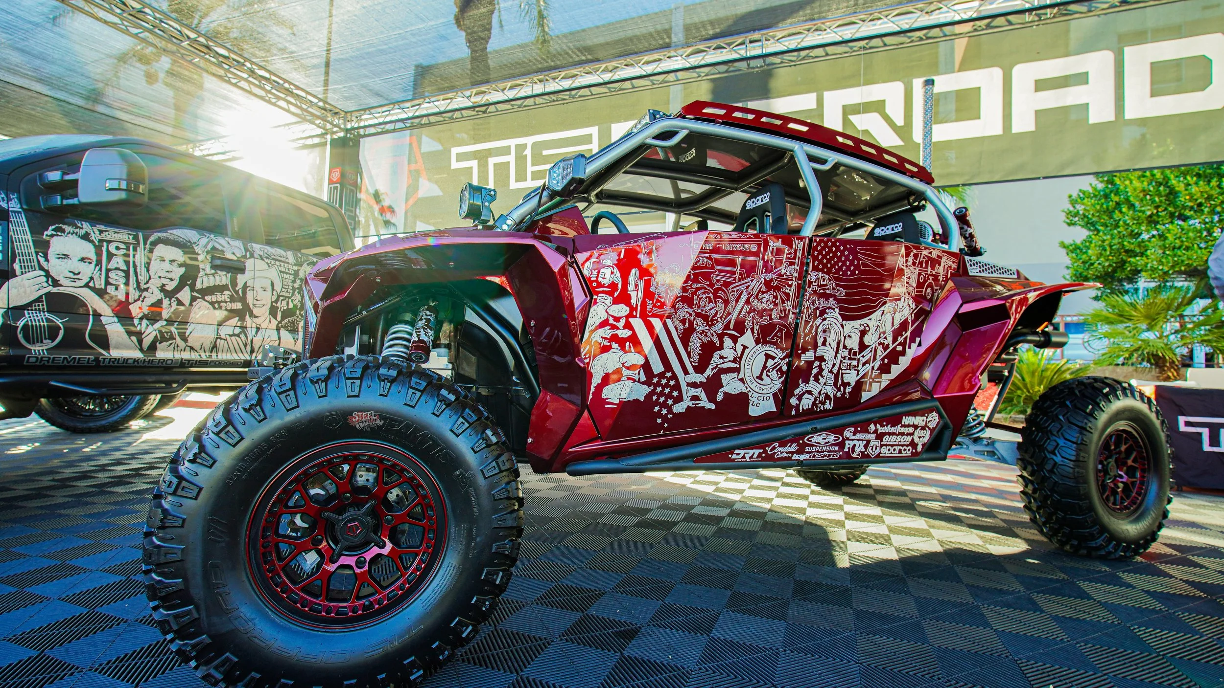 Custom off-road vehicle with red and black paint, large tires, and detailed graphic artwork on the sides, displayed at an outdoor event under a canopy with green plants in the background.
