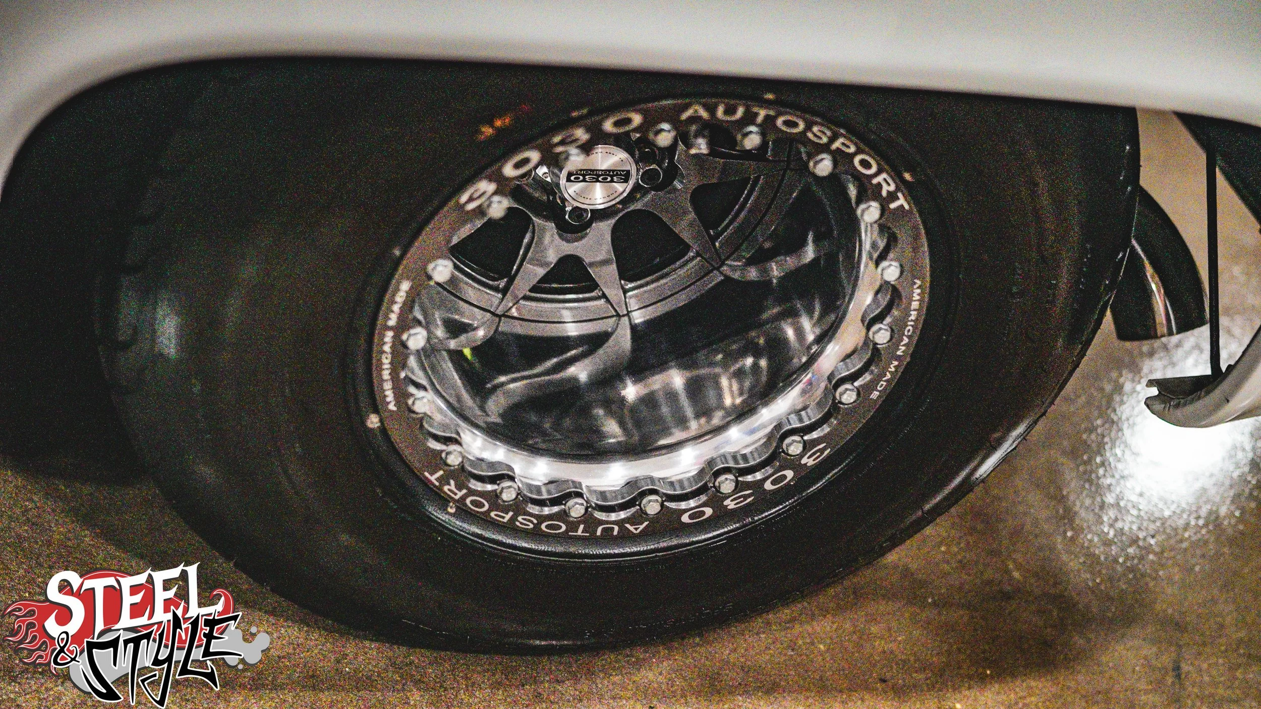 Close-up of a racing car tire with multi-compound tires and a detailed rim, showing the tire's sidewall and part of the car's body in the background.