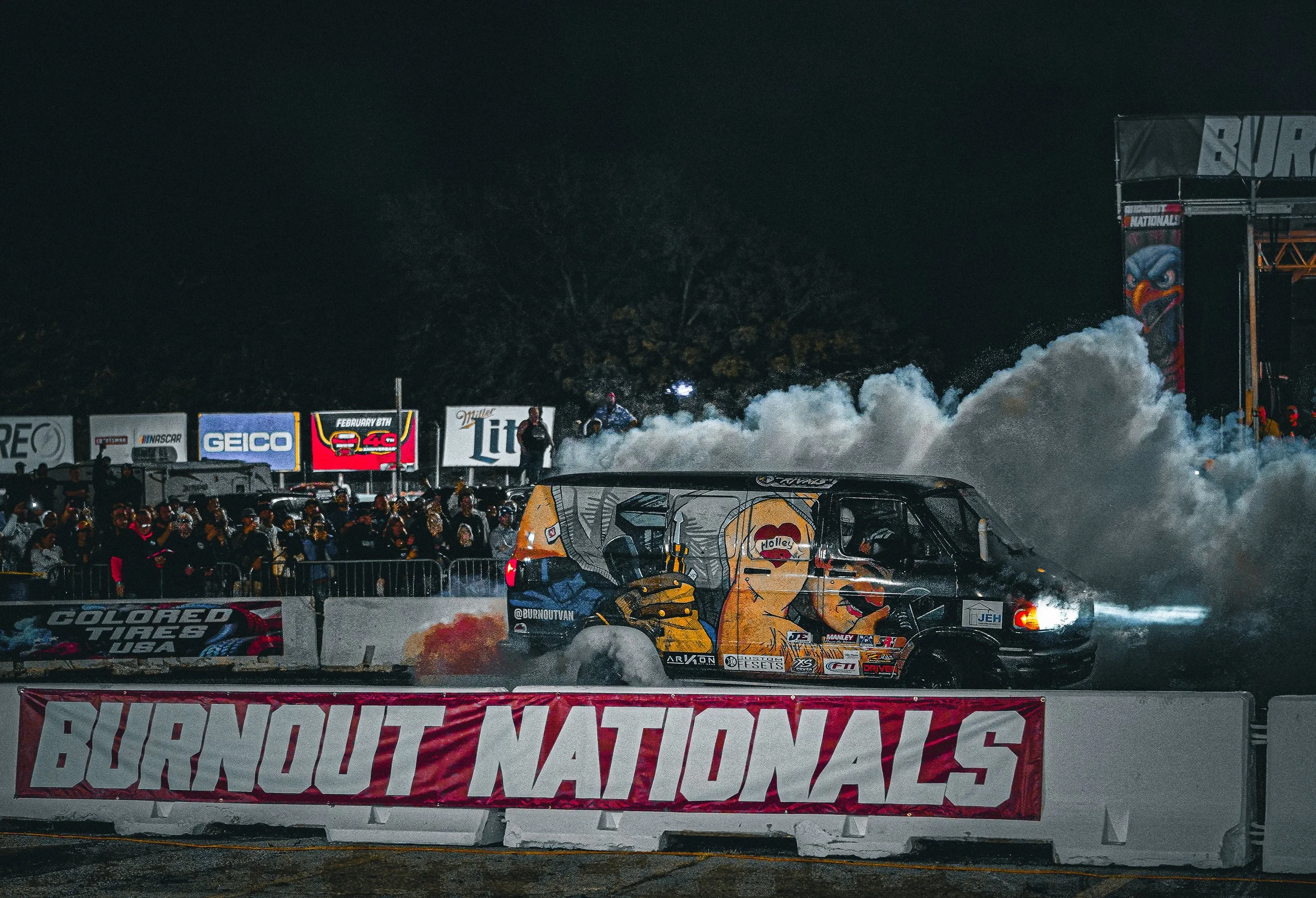 A van with colorful art and the logo 'Holley' on its side, performing a burnout at night during the Burnout Nationals event, with large clouds of smoke, an audience behind barriers, and various sponsor banners visible in the background.