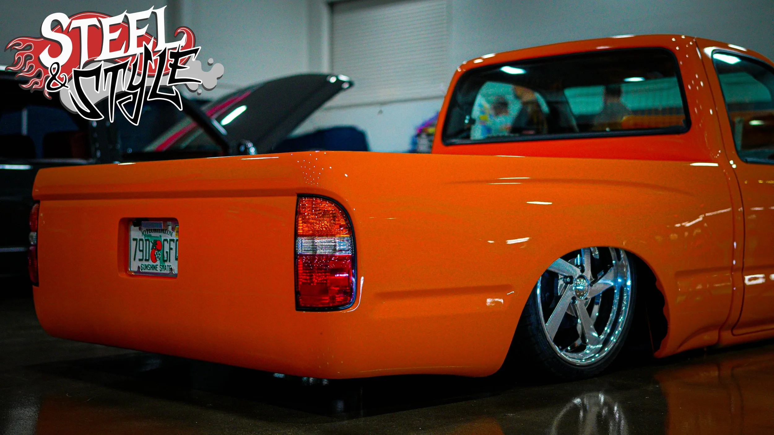 An orange lowered pickup truck with large chrome wheels and a reputable Florida license plate, parked indoors with other cars in the background and the words Steel & Style in graffiti-style text in the top left corner.