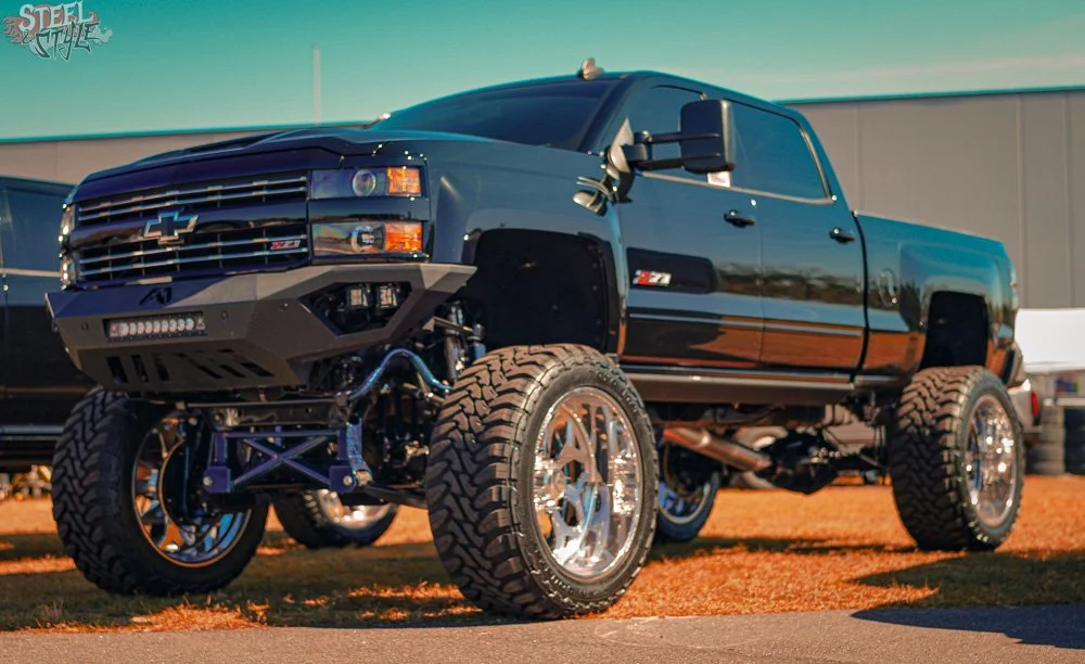 A black lifted Chevrolet Silverado pickup truck with large aftermarket wheels and tires, custom front bumper with LED lights, and a Z71 badge, parked on a grassy area with a building and other vehicles in the background.