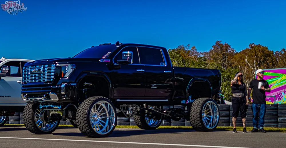 A highly customized black lifted pickup truck with large wheels and off-road tires parked at an outdoor event, with two people standing nearby.