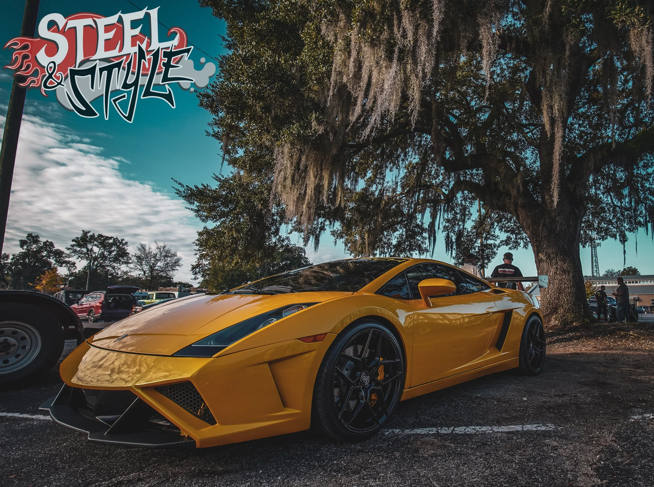Yellow Lamborghini sports car parked under a large tree at a car show with other vehicles and people in the background.