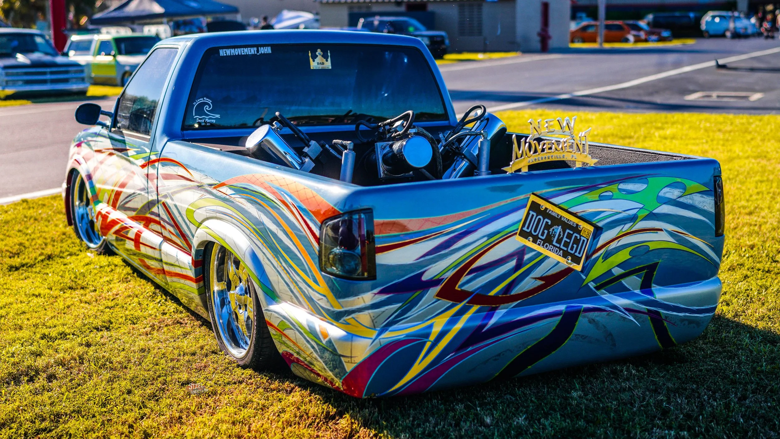 Colorful low-rider pickup truck with custom paint and a raised bed, parked on a grassy area at a car show