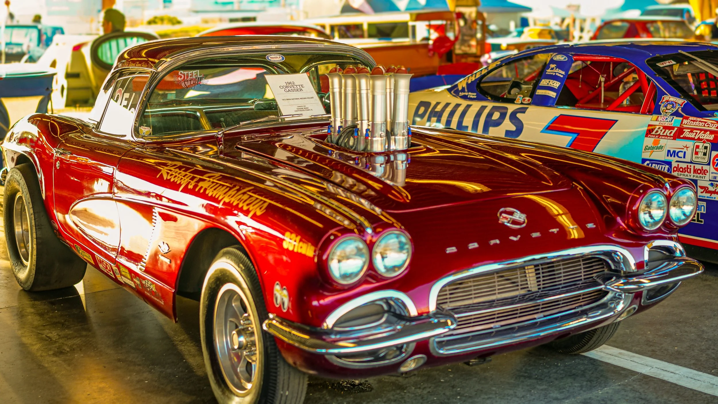 A red vintage Corvette race car with a large engine protruding from the hood, displayed at a car show with other race cars and vehicles in the background.