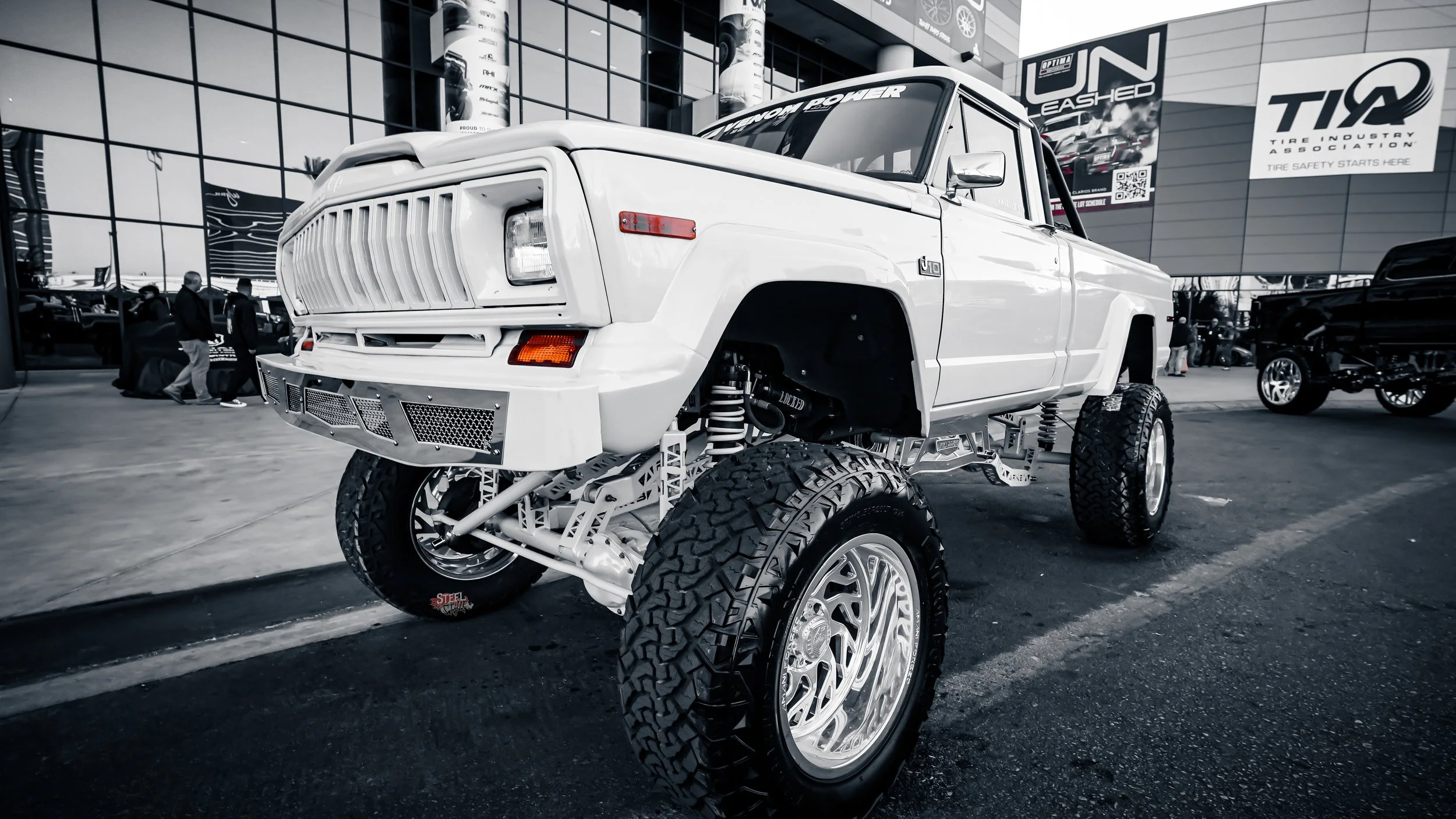 A modified white pickup truck with a lifted suspension, large off-road tires, and custom rim wheels parked outside a building with banners and posters on the windows, including the TIA Tire Industry Association logo.