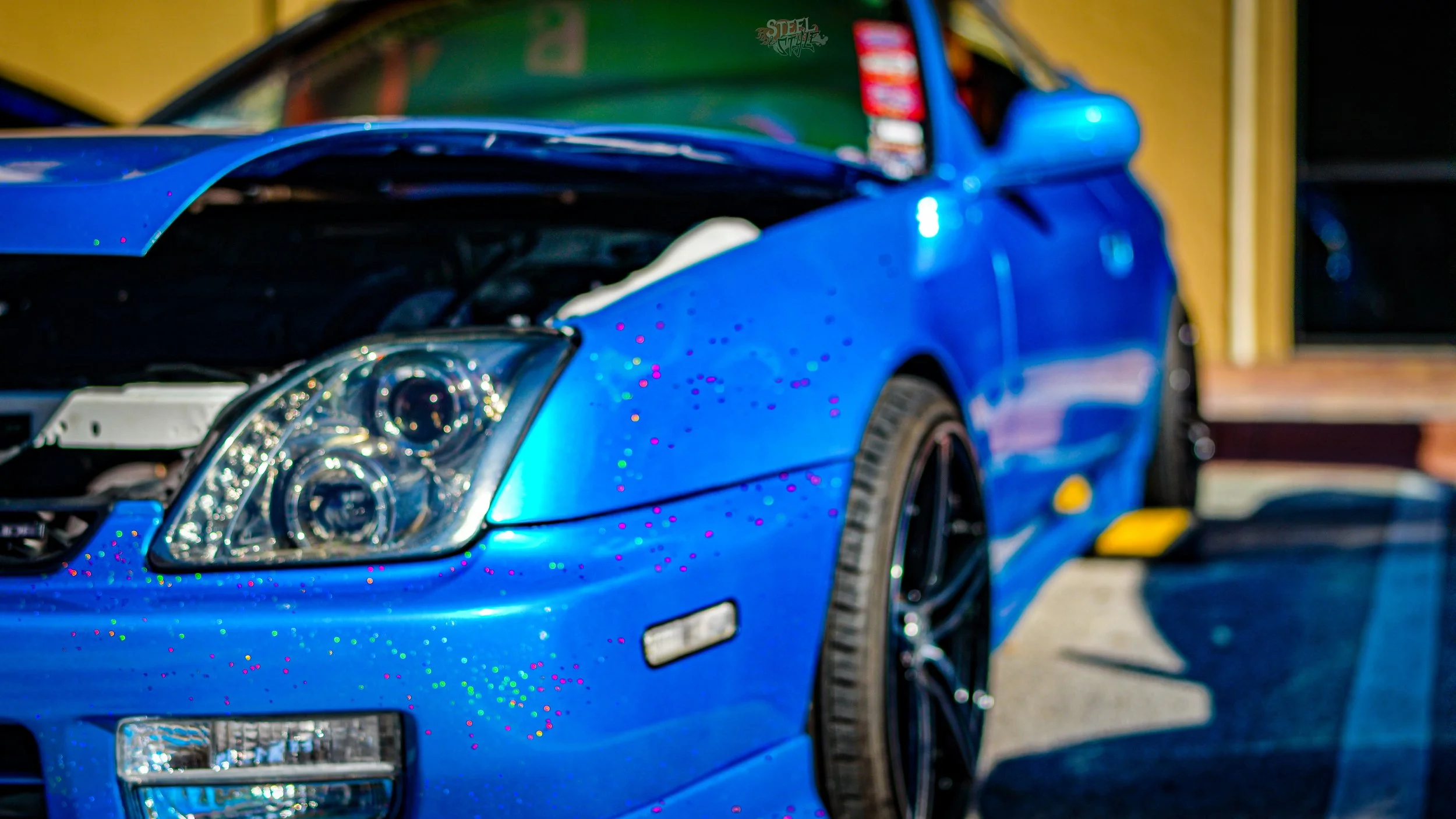 Close-up of a blue sports car with a partially open hood, showing the headlight and front wheel, parked outdoors on a sunny day.