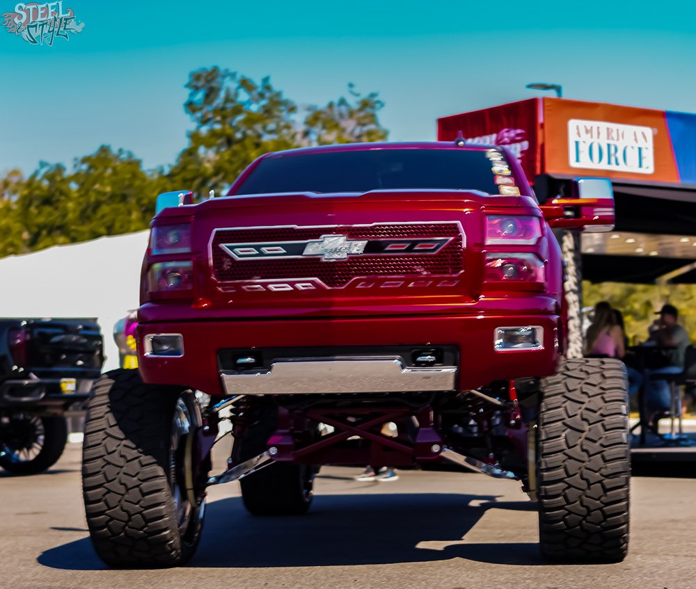 A red Chevrolet monster truck displayed outdoors, seen from the front, with large off-road tires and a custom grille.