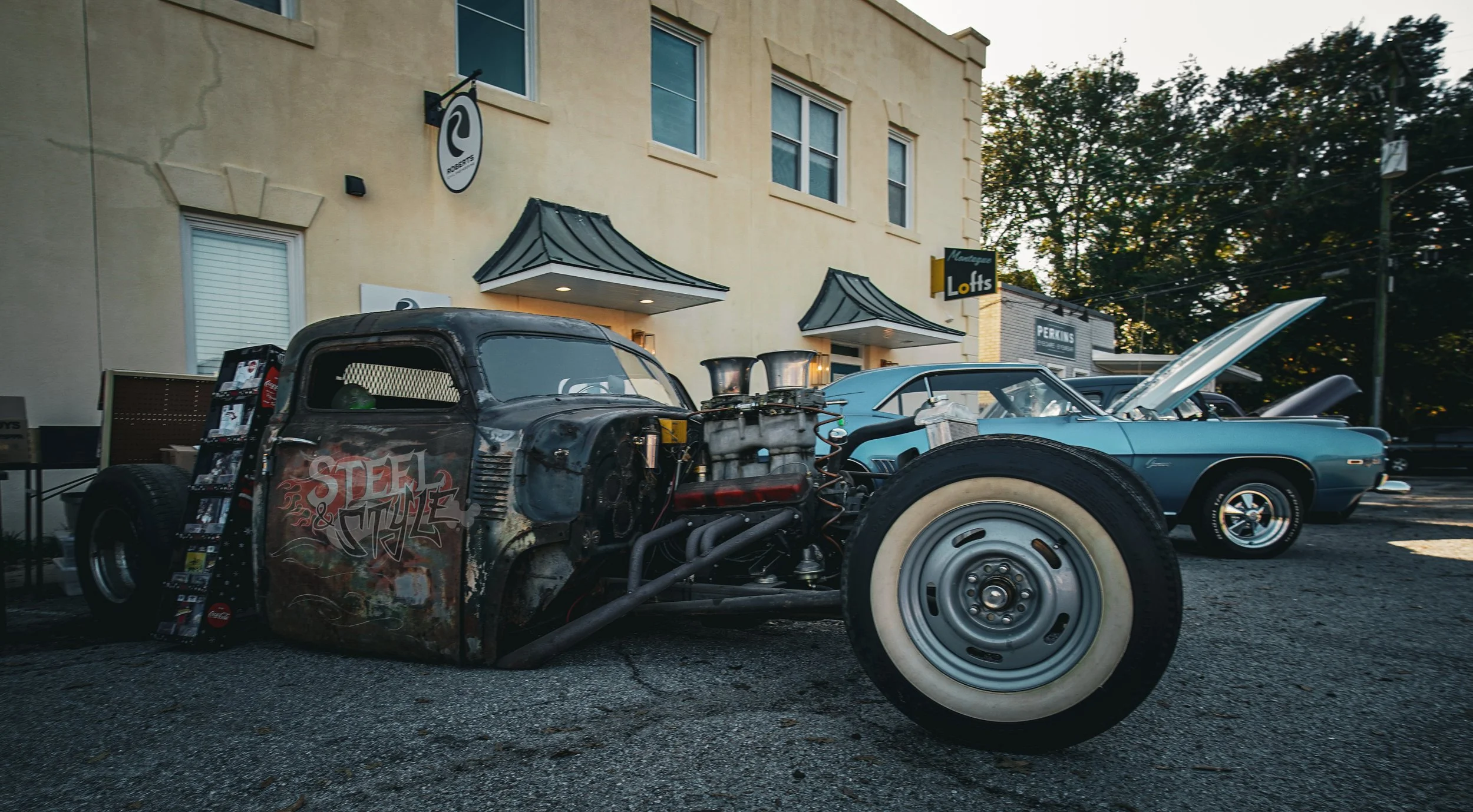 Old, weathered hot rod car with exposed engine and graffiti on the side parked in front of a building with vintage signs, alongside a classic light blue car with open hood and vintage chrome wheels. Trees are visible in the background.