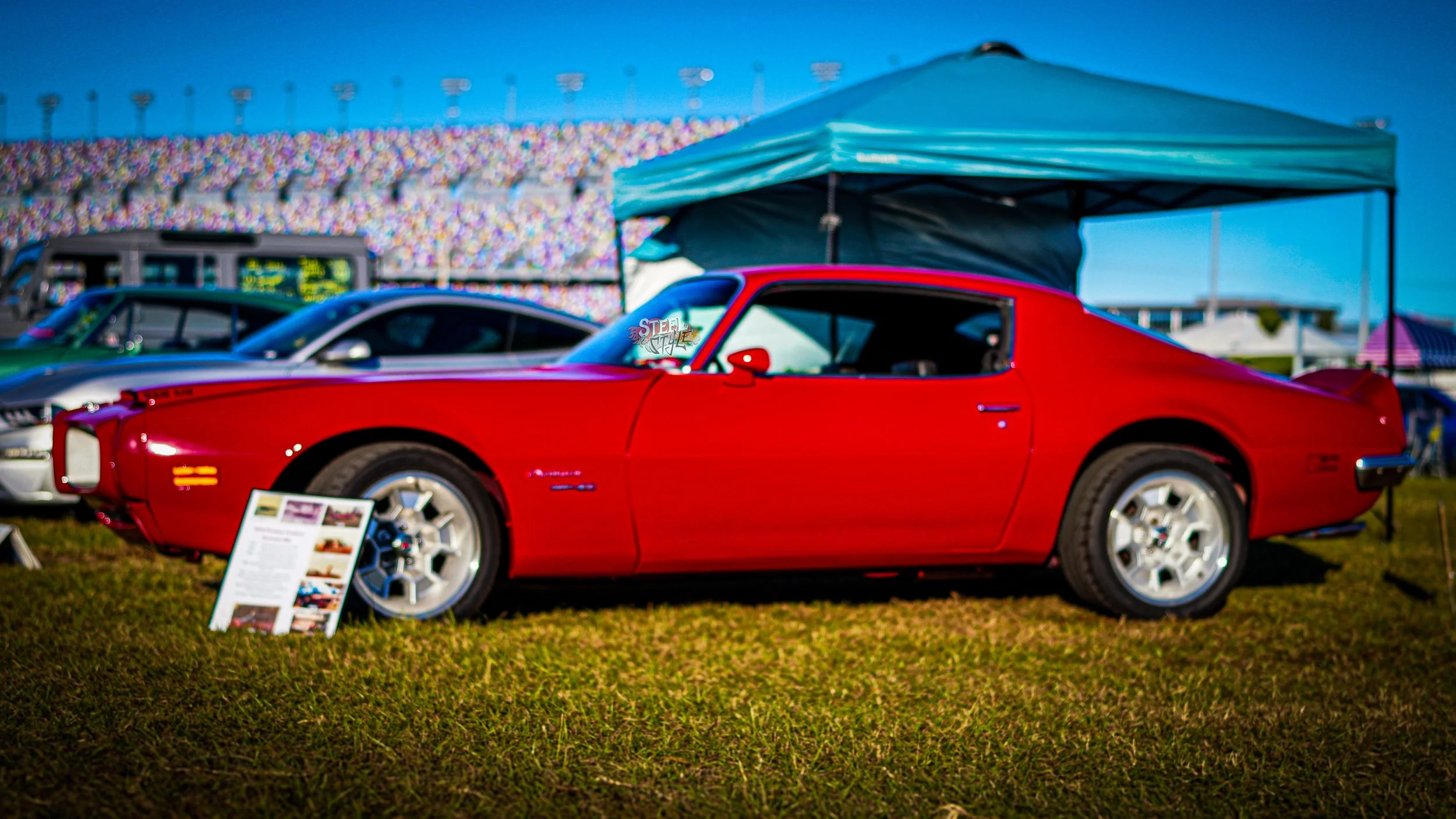 Red classic car displayed at a car show under a green canopy with other cars and a large grandstand in the background.