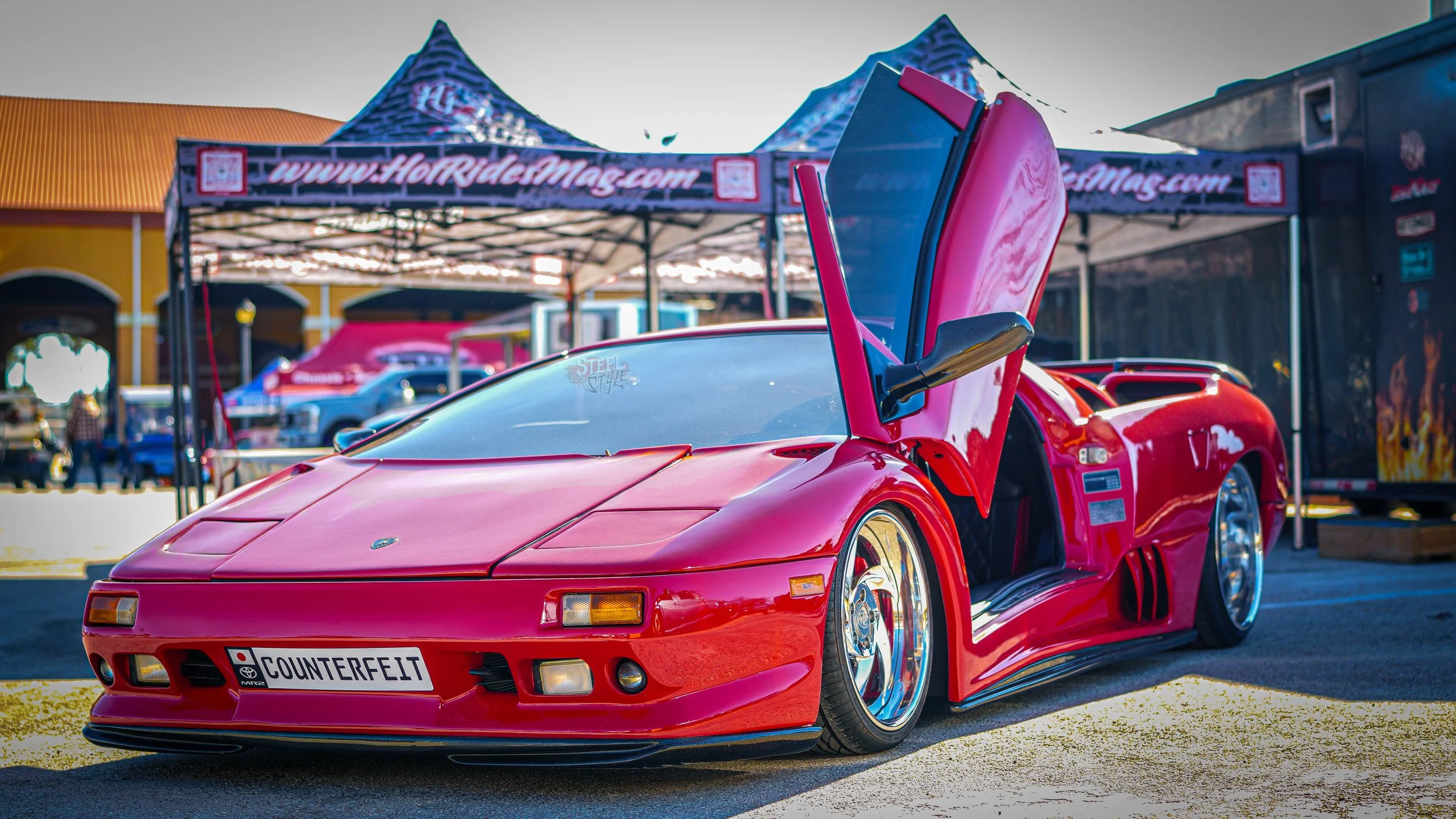 Red sports car with butterfly doors open at an outdoor car show, tents and people in the background.