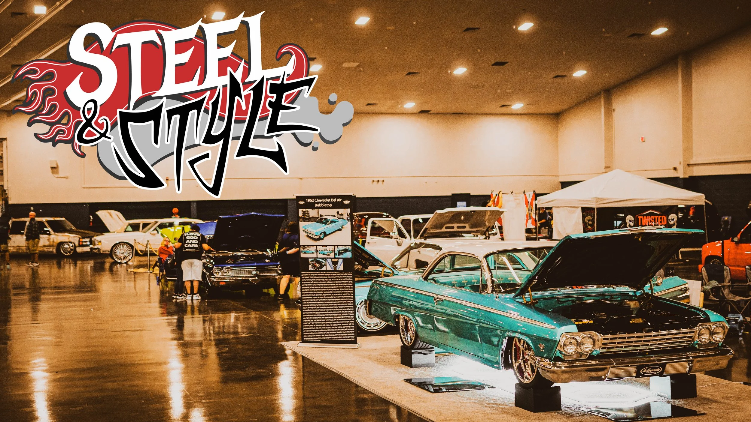 Classic cars on display at an indoor car show with a large graffiti style sign in the background that reads "Steel & Style".