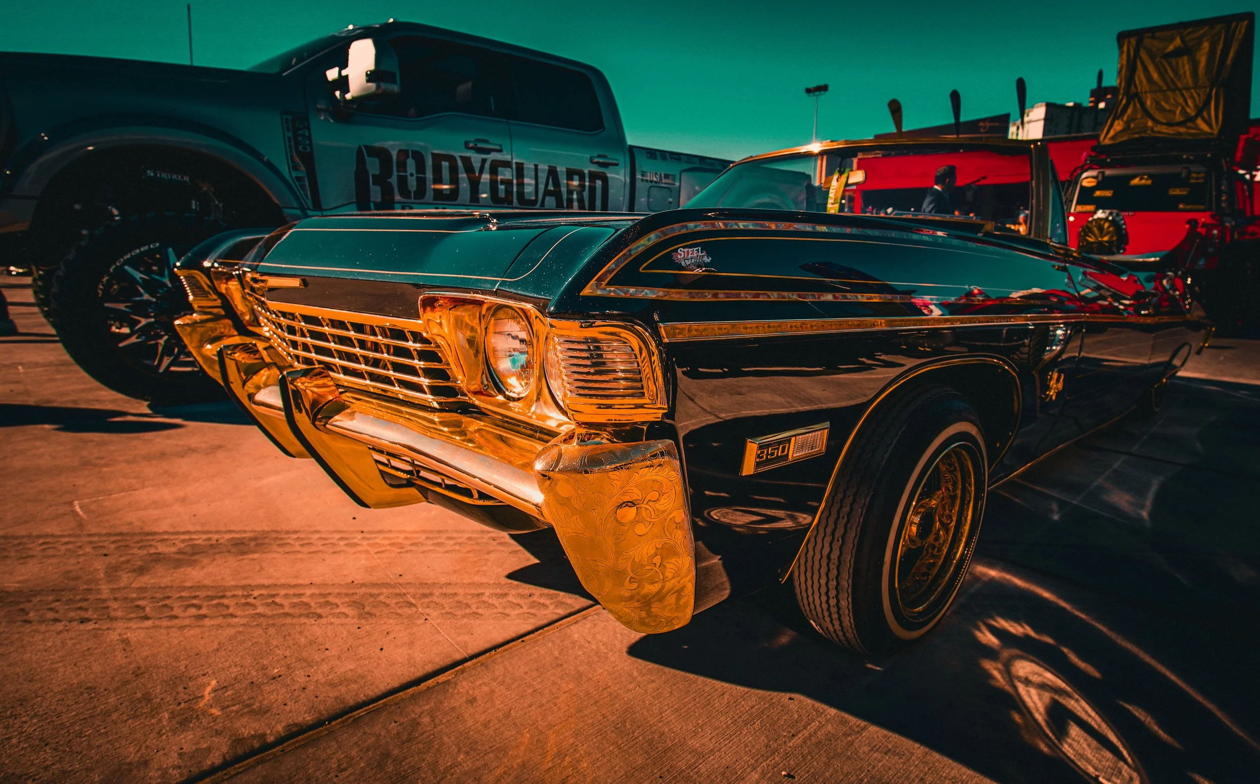 A vintage black and gold Pontiac Firebird car with '350' emblem on the side, parked on a concrete surface, with a large truck and other vehicles in the background under a clear sky.