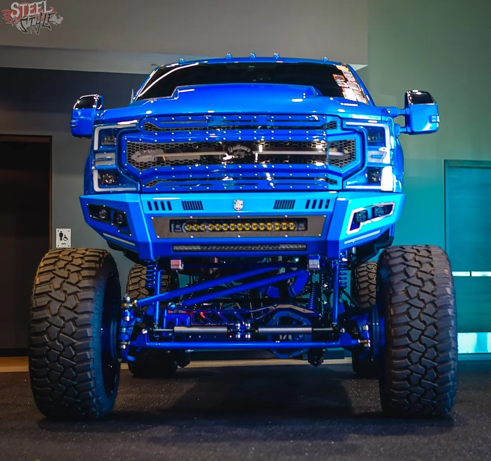 A custom blue lifted truck on display, showcasing its large tires, aggressive suspension, front grille, and lighting.
