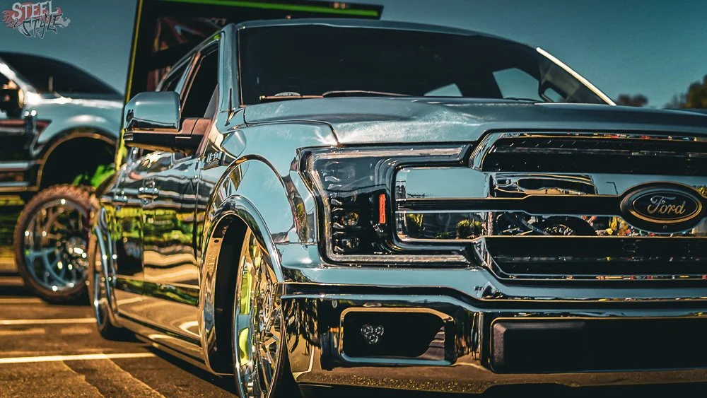A shiny, customized silver Ford truck on display at a car show, with other cars and a sign in the background.