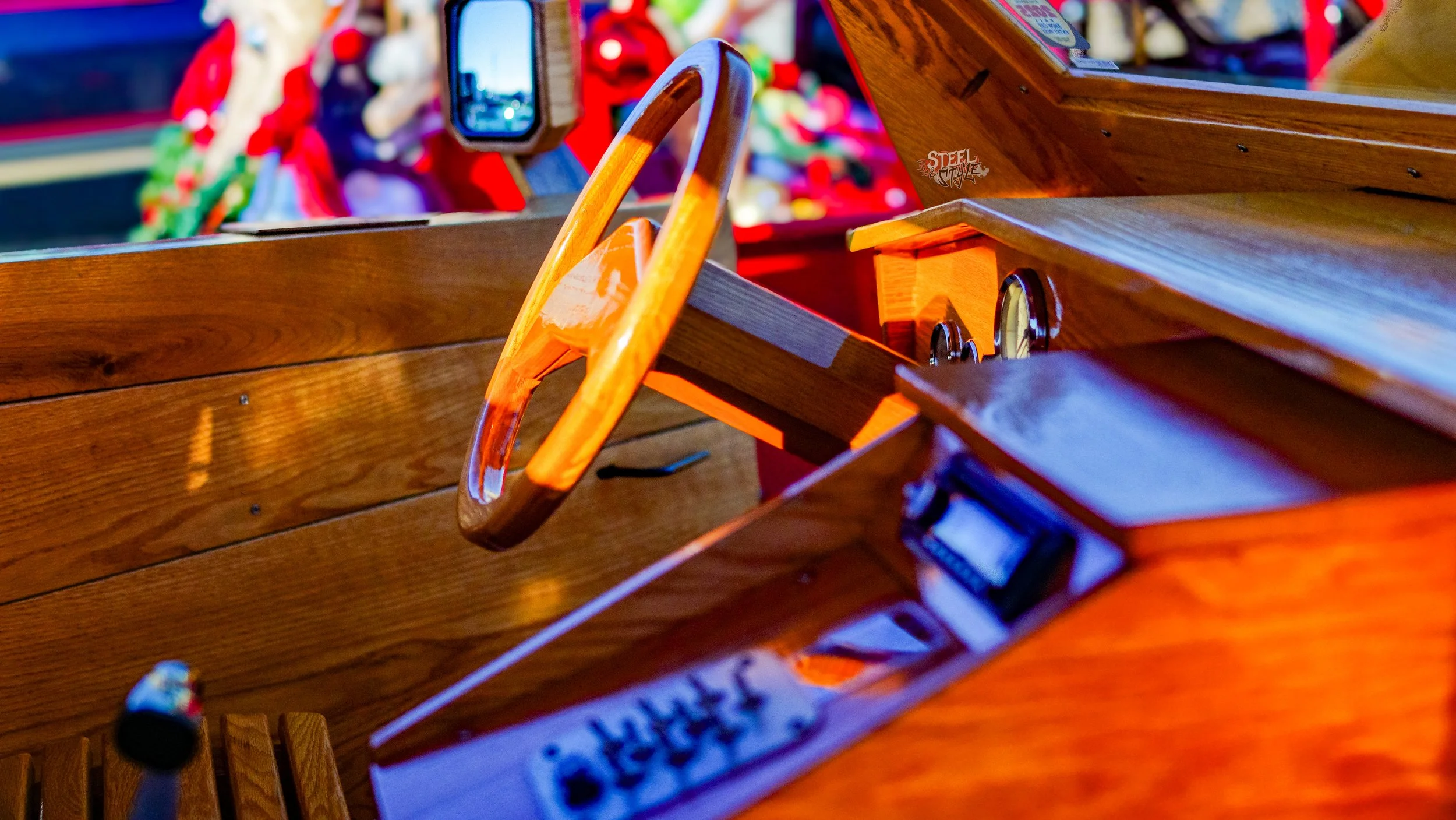 Close-up image of a wooden vintage pedal car dashboard with a steering wheel and an open compartment, with colorful objects blurred in the background.