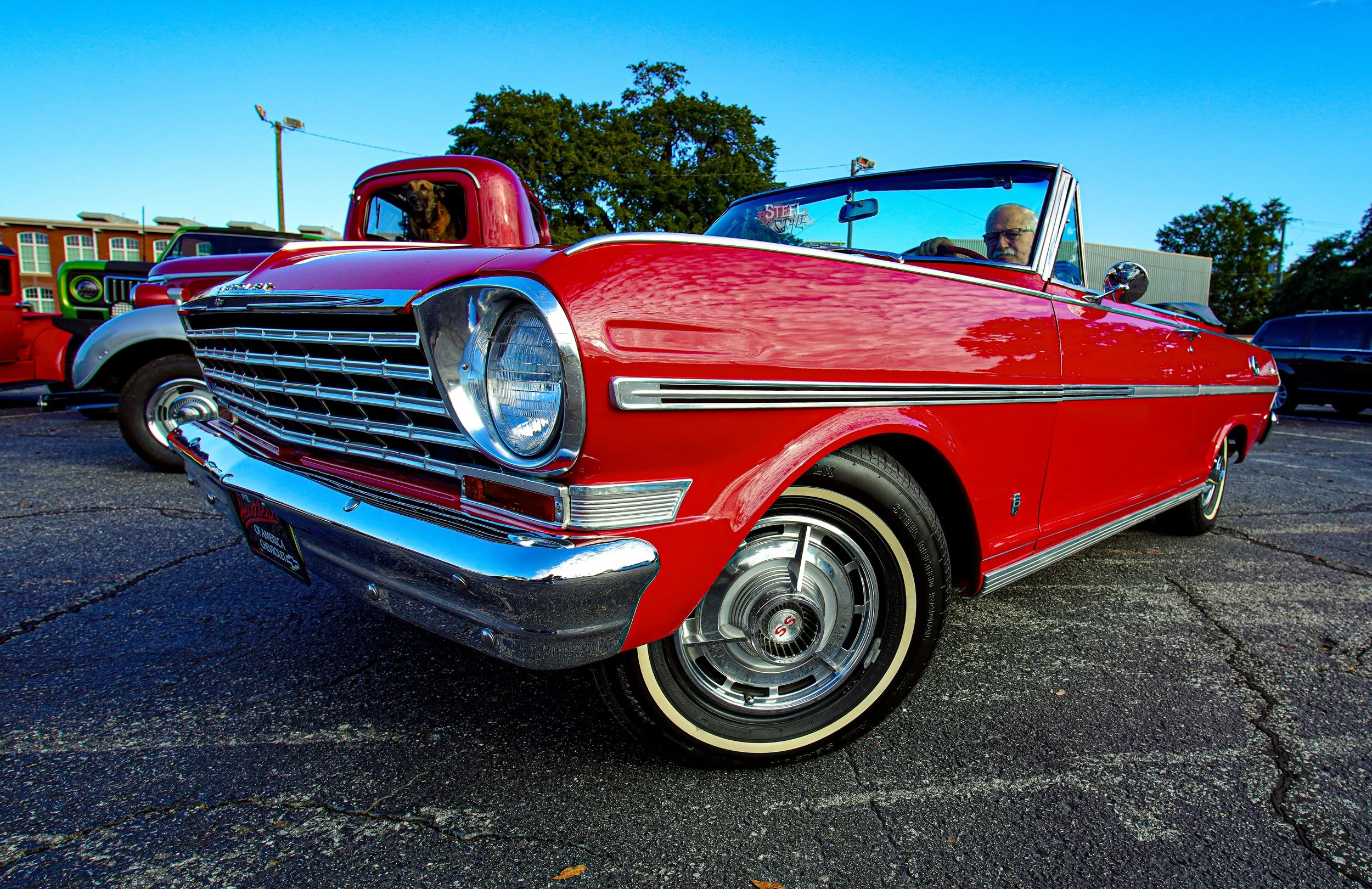 A classic red convertible car at a car show, with a man driving and a dog in the passenger seat, with other vintage cars in the background and a clear blue sky overhead.
