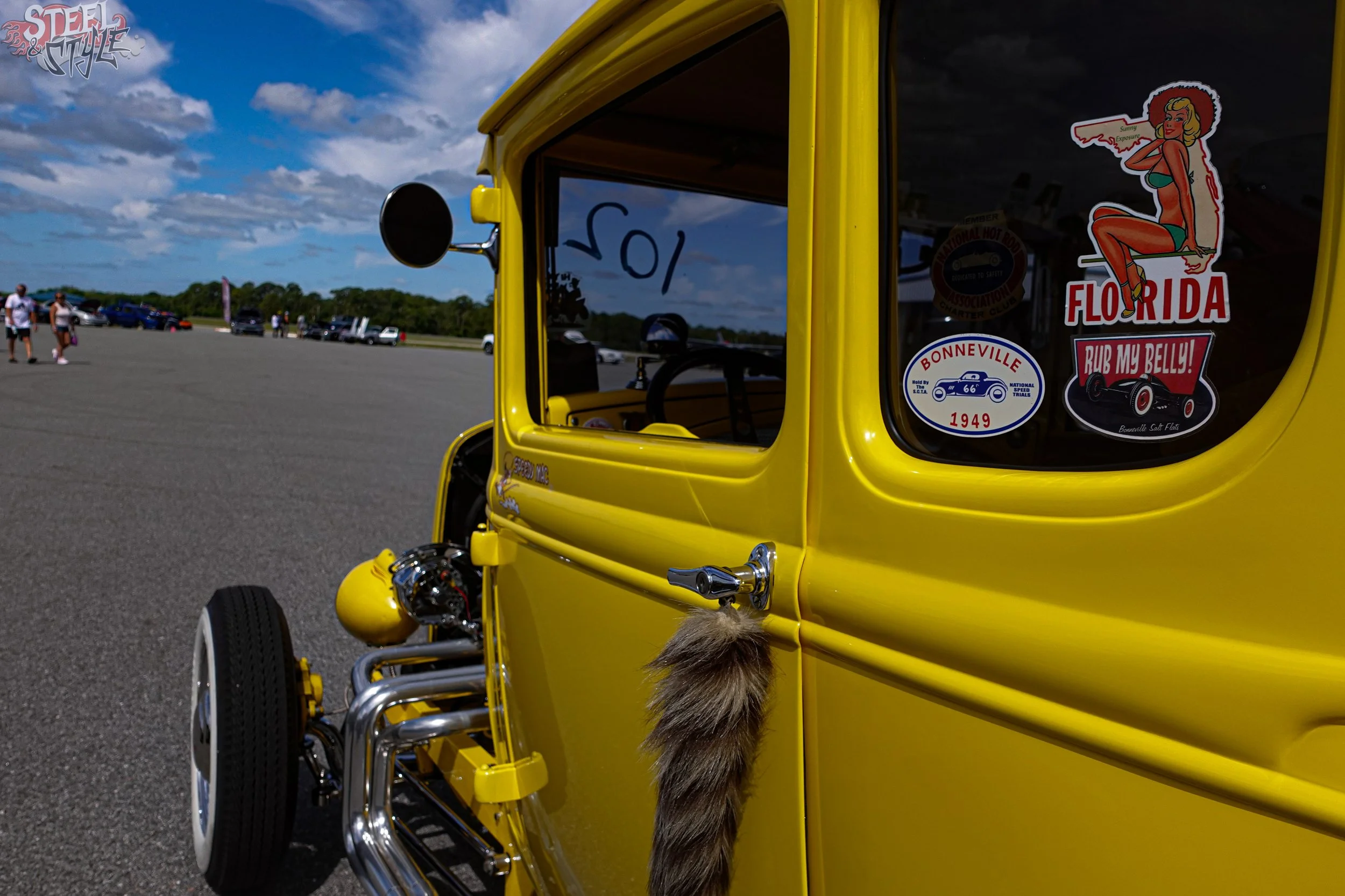 Yellow vintage hot rod car with stickers on the window, parked in an outdoor lot with people and other cars in the background, under partly cloudy sky.