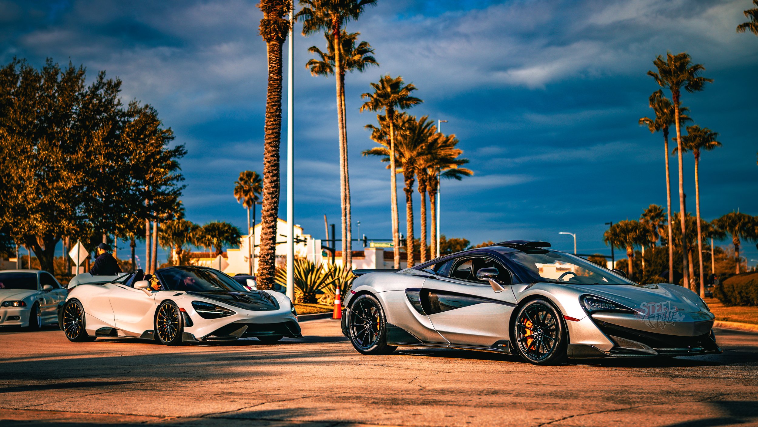 Two silver and black luxury sports cars parked on a street lined with palm trees under an orange and blue sky.