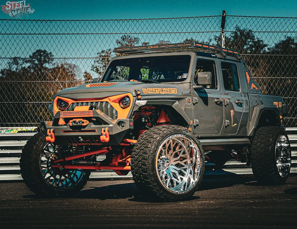 A customized off-road pickup truck with large custom wheels, lifted suspension, and aggressive design elements, parked on a racetrack with fencing and trees in the background.