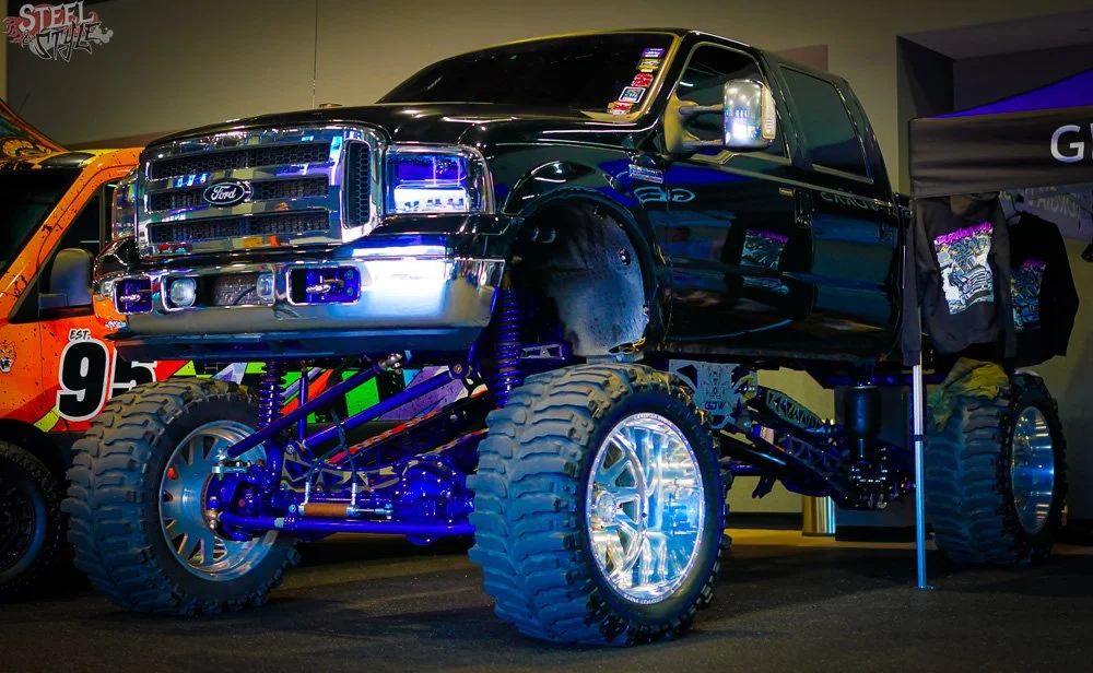 A custom lifted Ford truck with large off-road tires, chrome wheels, and a shiny black body on display at an indoor event.