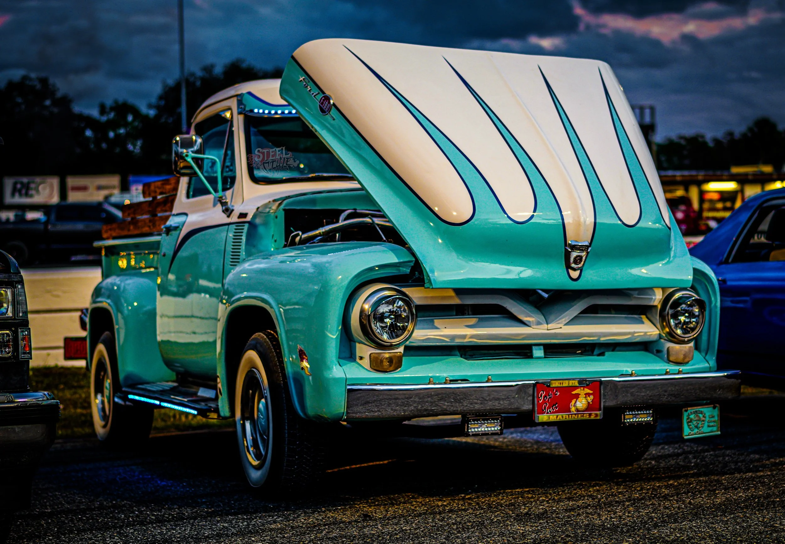 A vintage turquoise pickup truck with a white hood and black stripes, parked at night with its hood open. The truck has decorative blue LED lights underneath and vintage-style headlights. There are other vehicles parked nearby and a cloudy sky in the